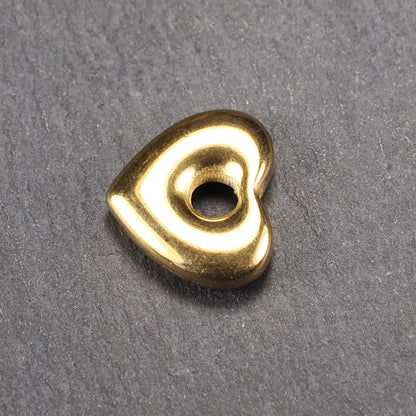 Goldfarbenes, hochglänzendes, abgerundetes Herz mit rundem mittigem Loch, glatte Oberfläche, auf dunkel strukturiertem Untergrund.