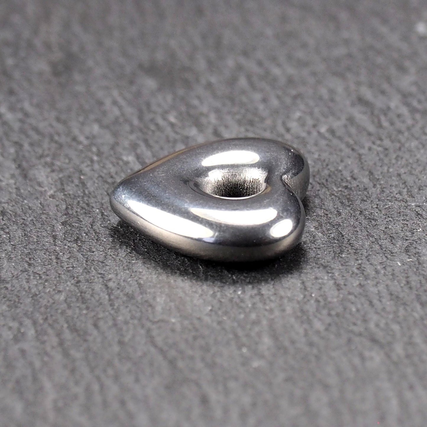 Herzförmiger, silberfarben glänzender Metallanhänger mit glatter Oberfläche und mittigem rundem Fädelloch.