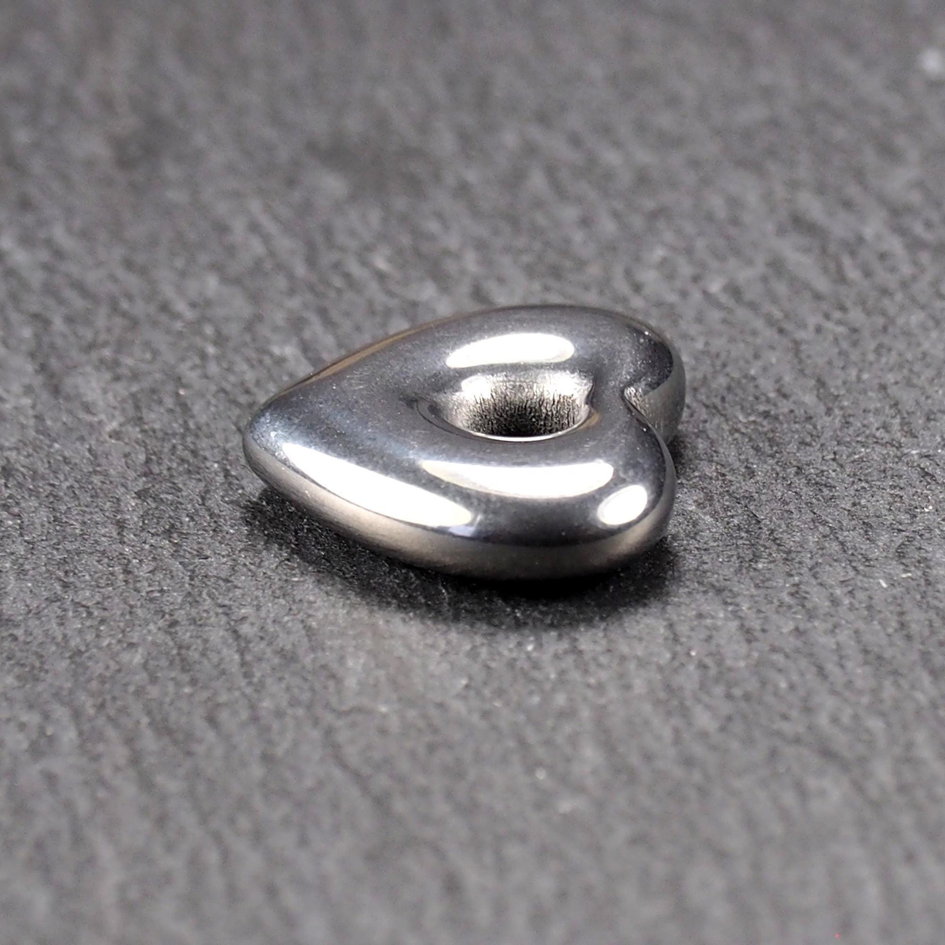 Herzförmiger, silberfarben glänzender Metallanhänger mit glatter Oberfläche und mittigem rundem Fädelloch.