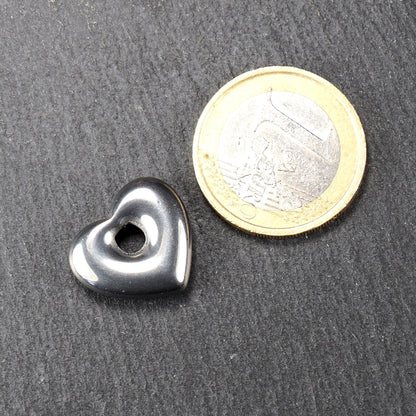 Herzförmiger Anhänger, metallisch dunkelgrau, glänzend poliert, mit rundem mittigem Fädelloch; daneben eine Ein-Euro-Münze zum Größenvergleich.