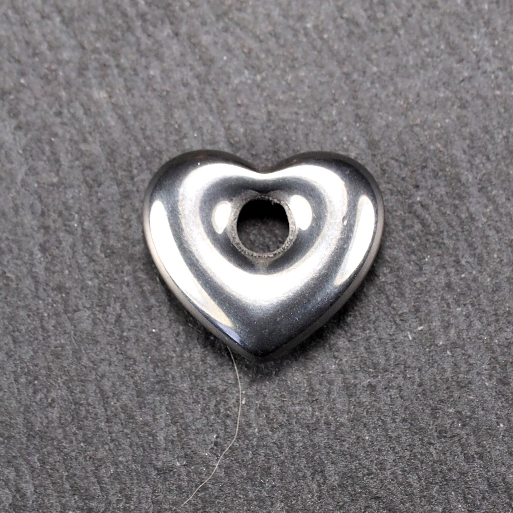 Herzförmiger, silberfarben glänzender Metallanhänger mit glatter Oberfläche und mittigem rundem Fädelloch.