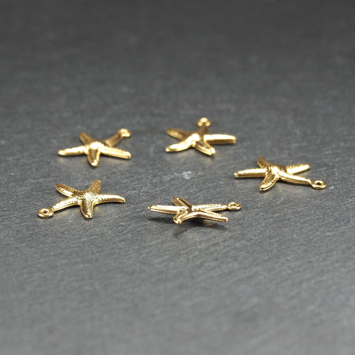 Fünf goldfarbene, flache Seestern-Anhänger mit reliefartiger, strukturierter und glänzender Oberfläche, jeweils mit kleiner runder Öse zum Aufhängen.