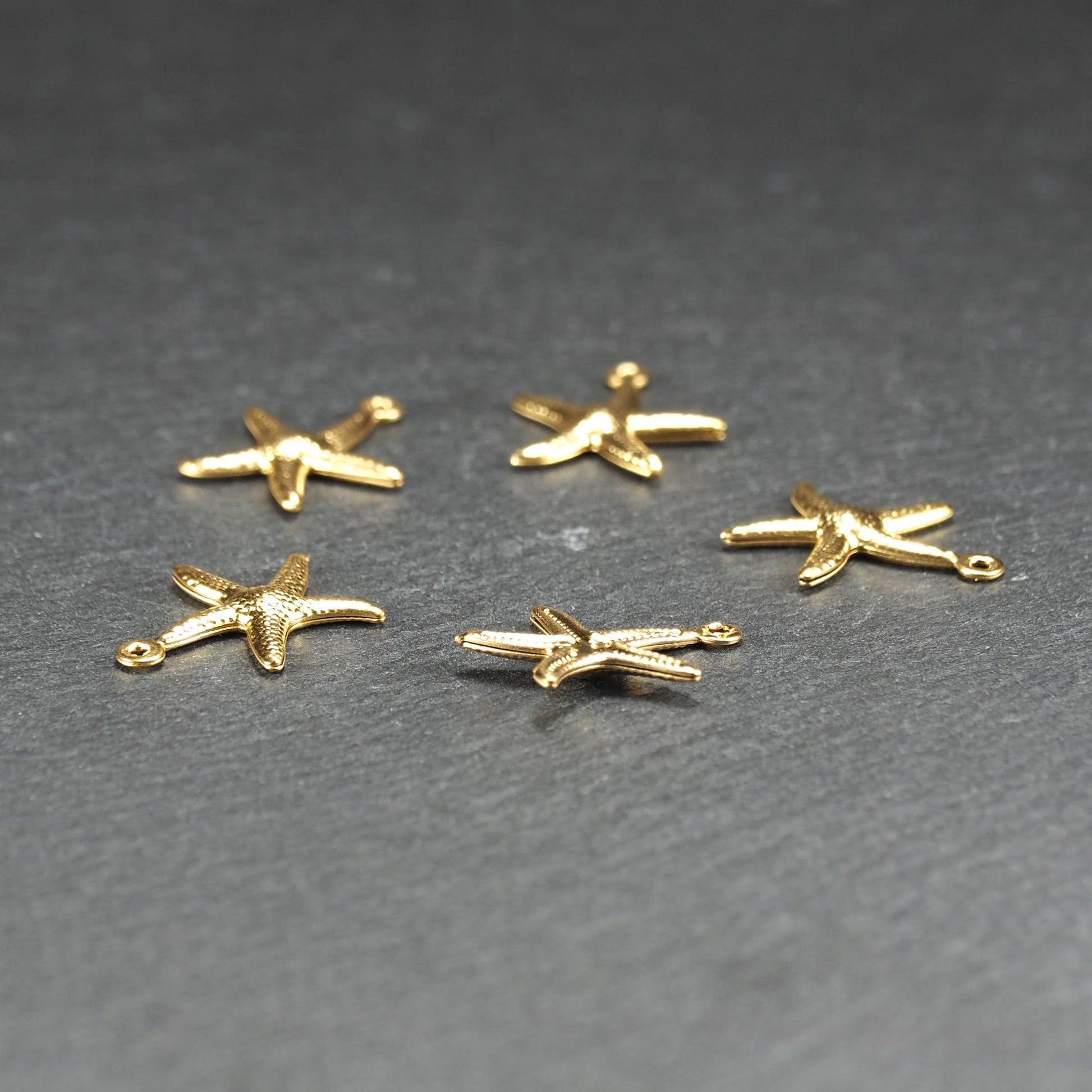 Fünf goldfarbene, flache Seestern-Anhänger mit reliefartiger, strukturierter und glänzender Oberfläche, jeweils mit kleiner runder Öse zum Aufhängen.