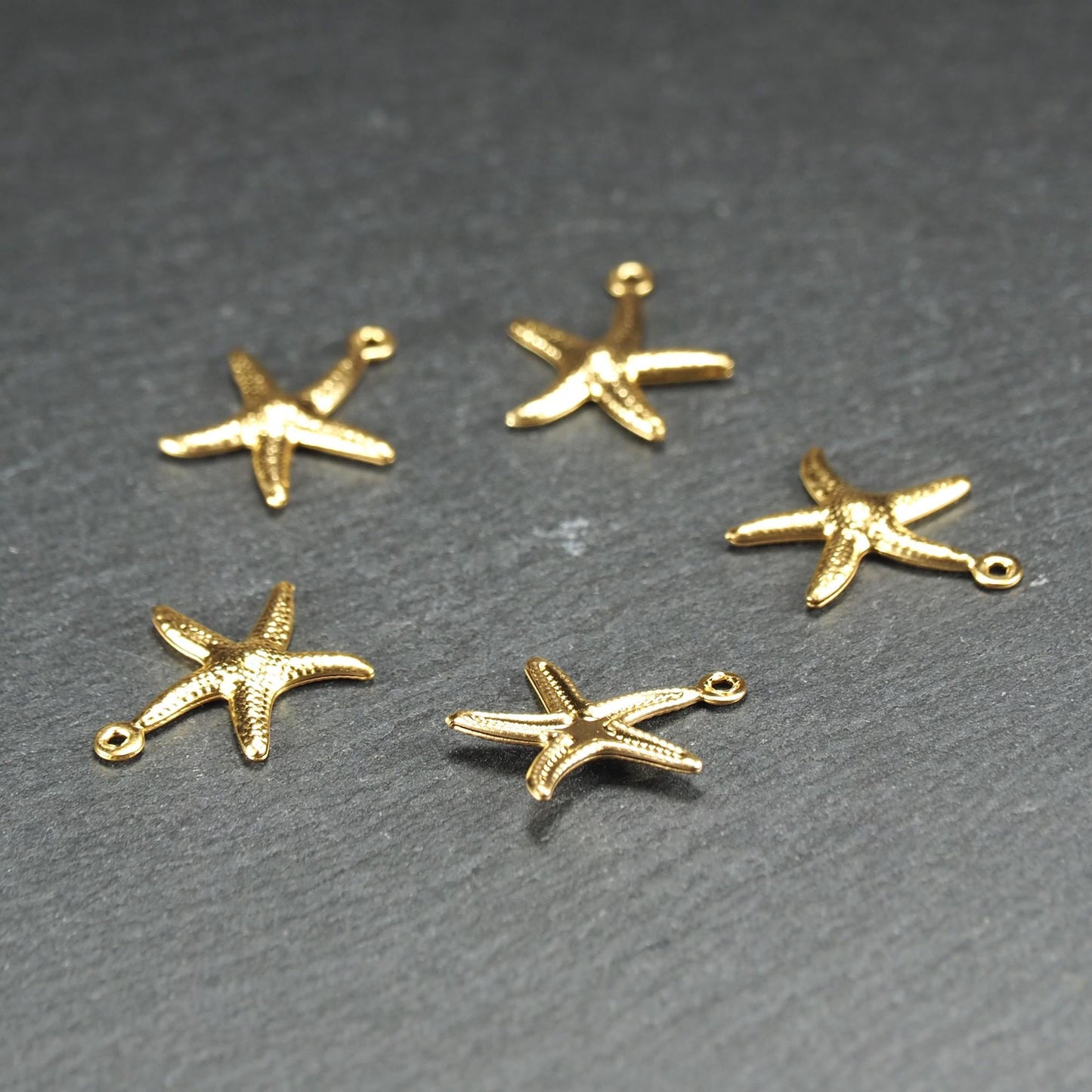 Fünf goldfarbene, flache Seestern-Anhänger mit reliefartiger, strukturierter und glänzender Oberfläche, jeweils mit kleiner runder Öse zum Aufhängen.