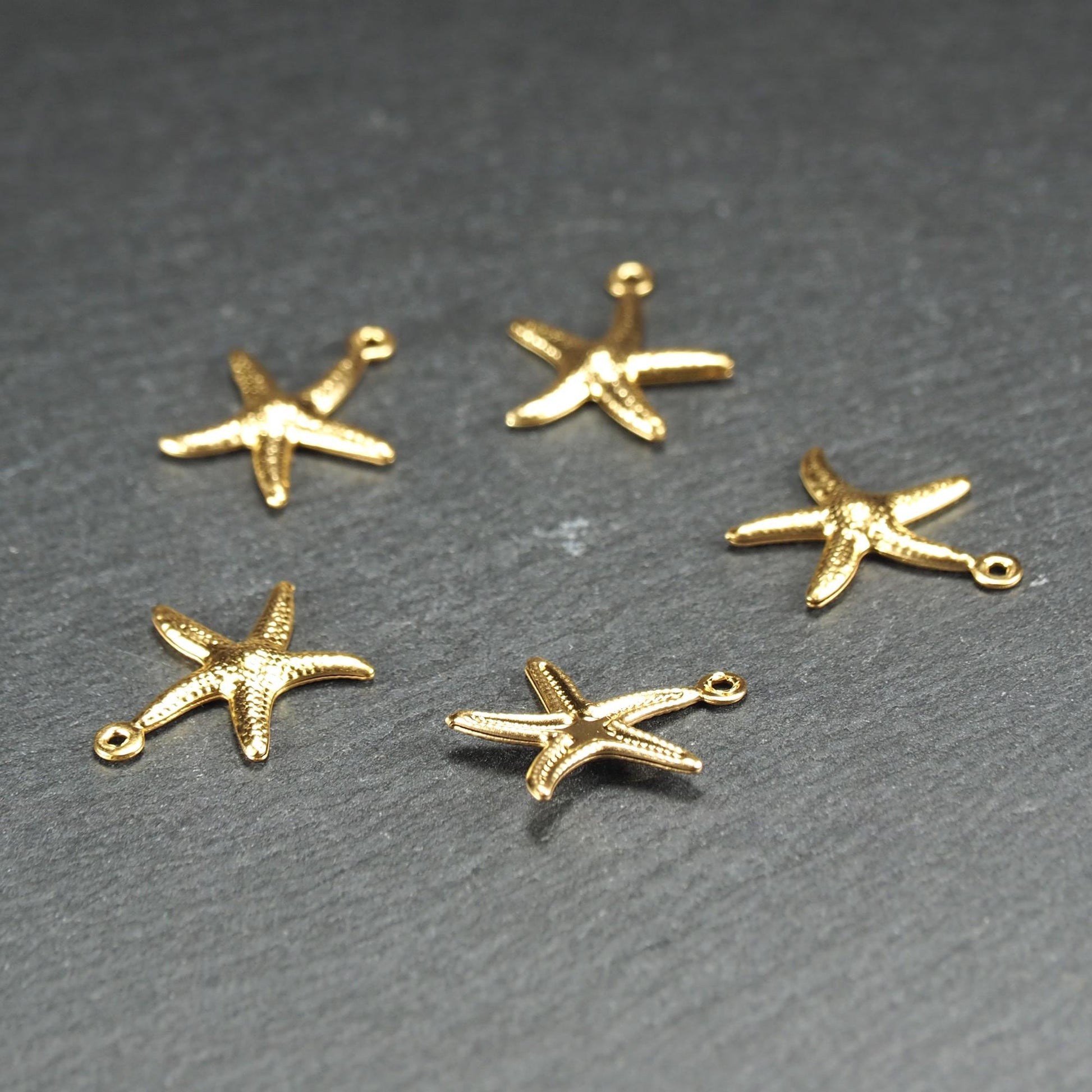 Fünf goldfarbene, flache Seestern-Anhänger mit reliefartiger, strukturierter und glänzender Oberfläche, jeweils mit kleiner runder Öse zum Aufhängen.