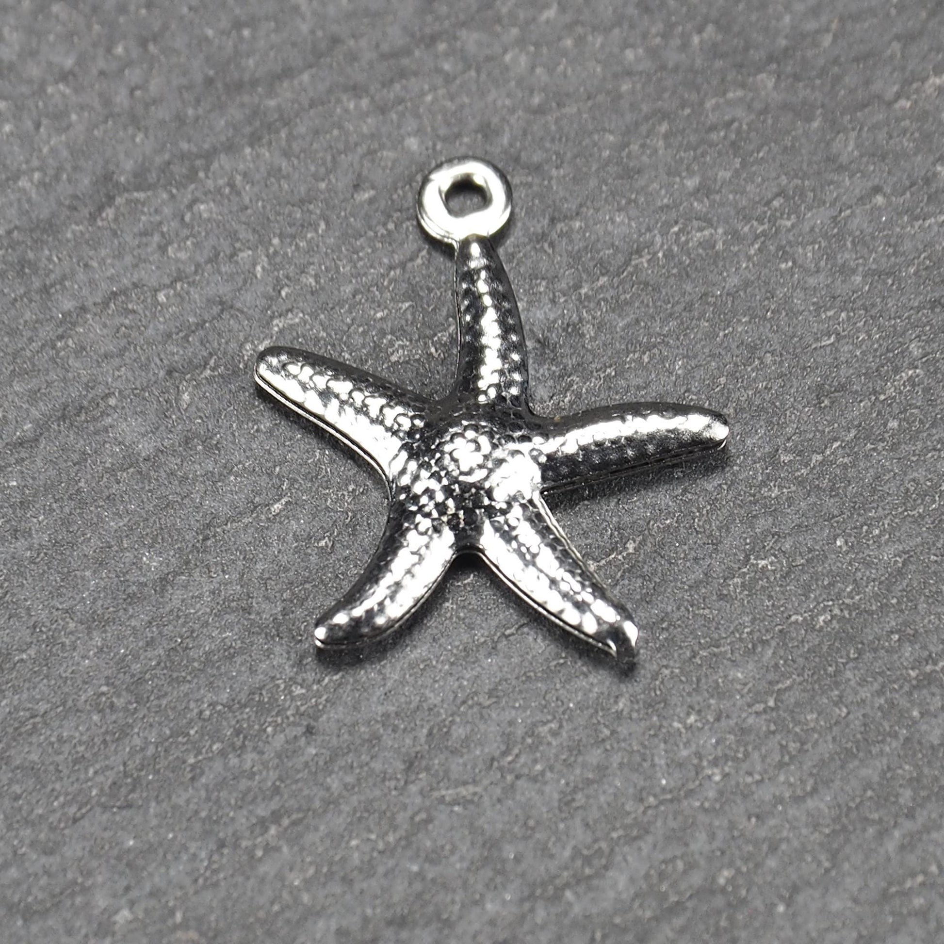 Seestern-Anhänger aus silberfarbenem Metall mit reliefartiger, strukturierter glänzender Oberfläche, fünf Armen und kleiner runder Öse oben.