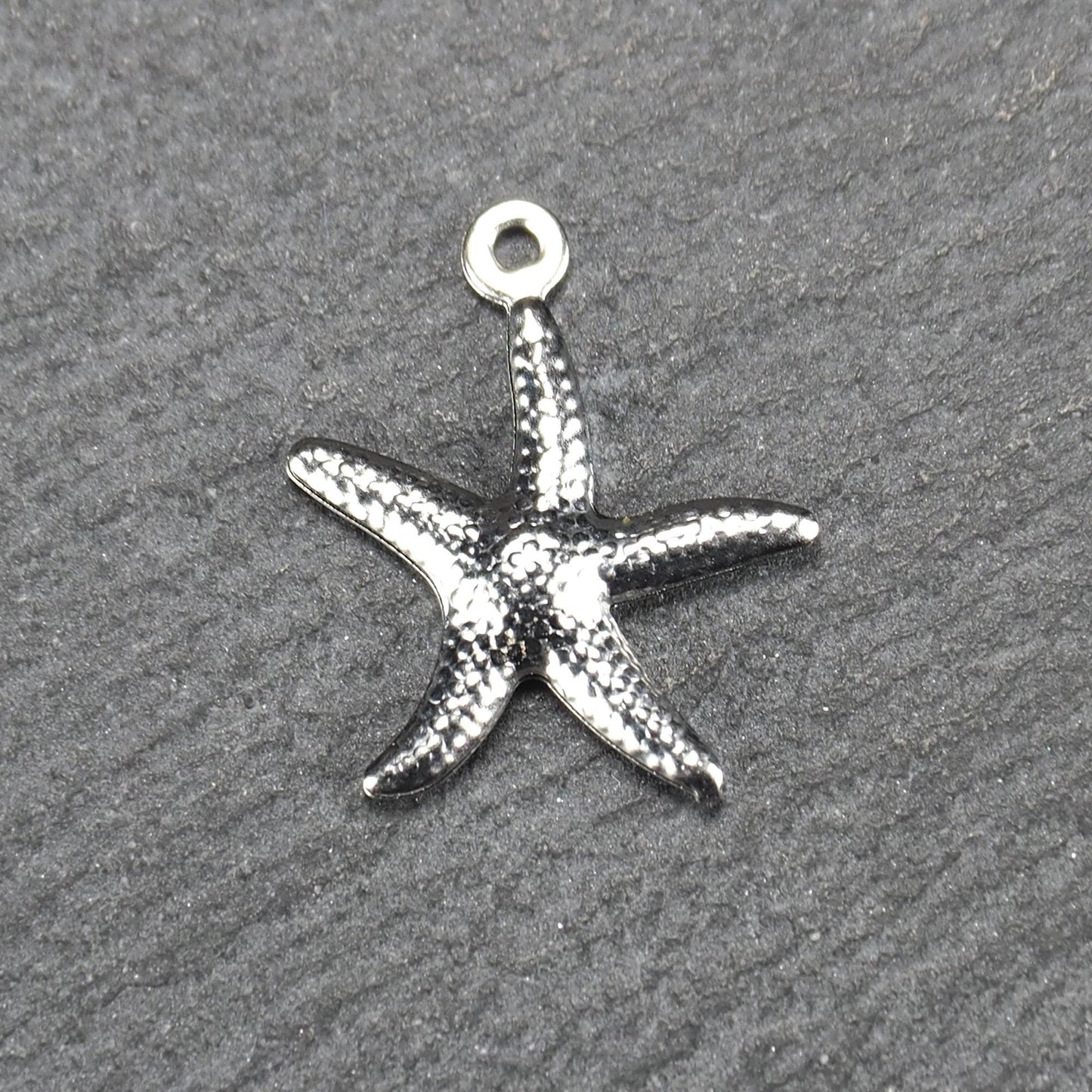 Seestern-Anhänger aus silberfarbenem Metall mit reliefartiger, strukturierter glänzender Oberfläche, fünf Armen und kleiner runder Öse oben.