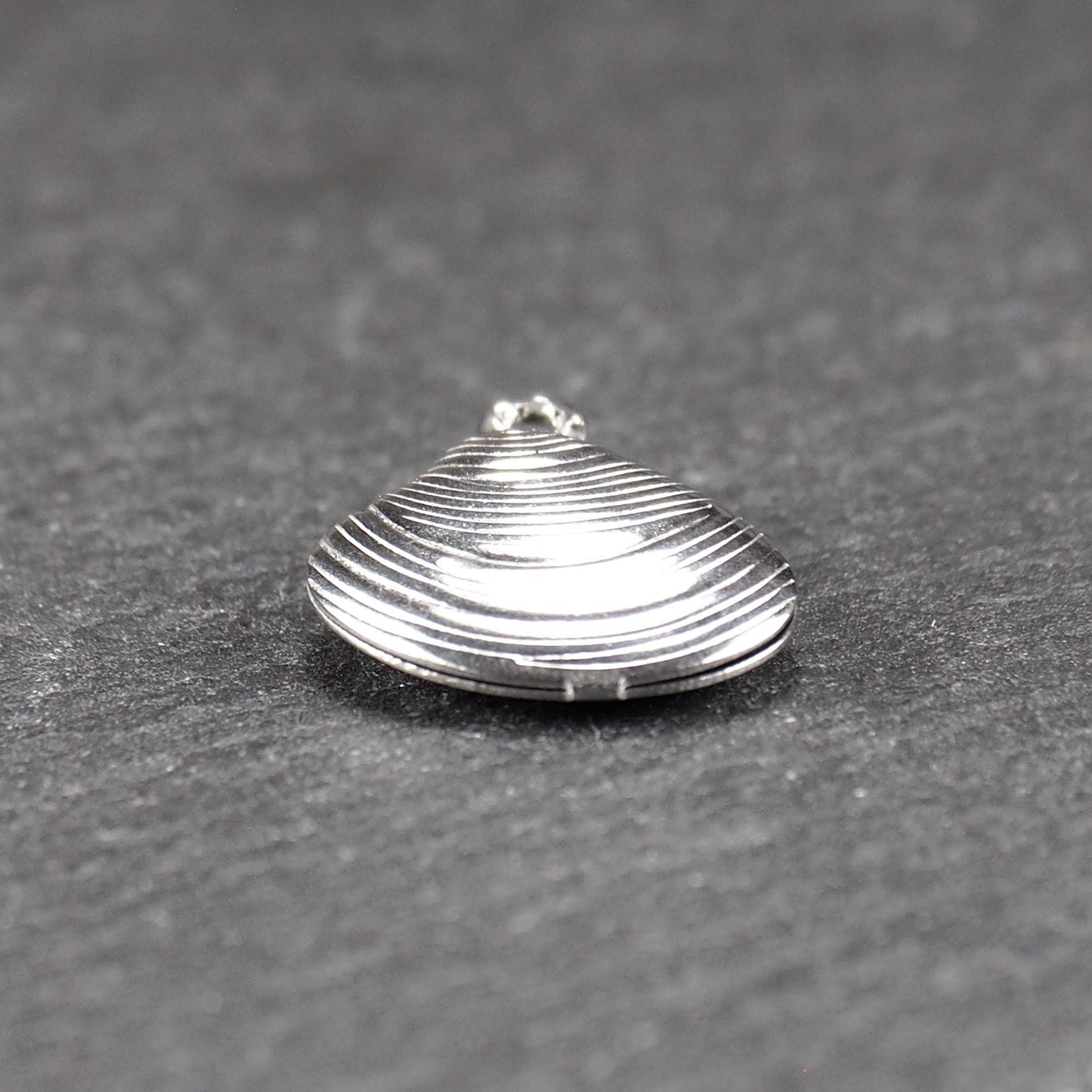 Muschelförmiger silberfarbener Anhänger mit polierter, glänzender Oberfläche, konzentrisch geriffelten Rillen und integrierter runder Öse oben.