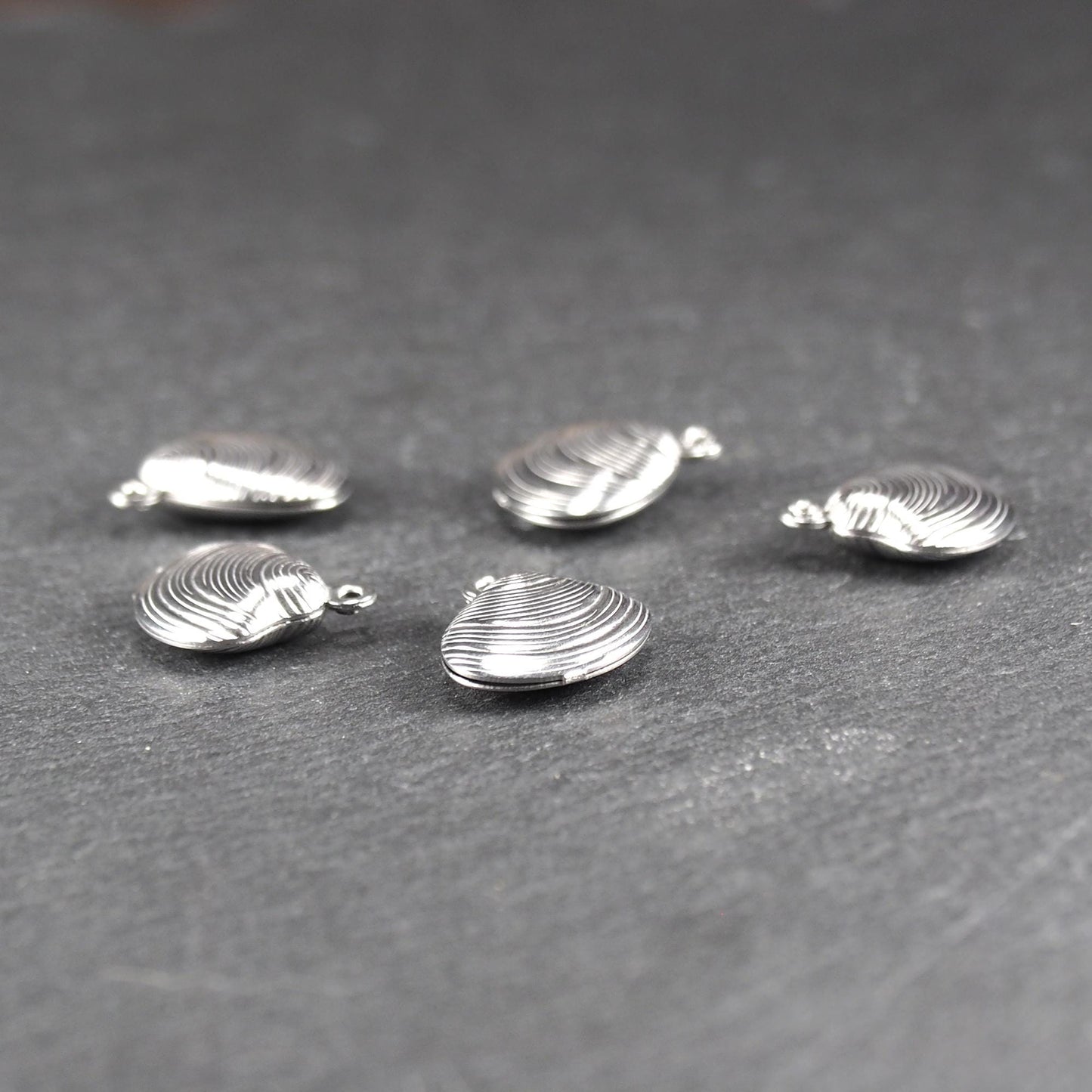 Muschelförmiger silberfarbener Anhänger mit polierter, glänzender Oberfläche, konzentrisch geriffelten Rillen und integrierter runder Öse oben.