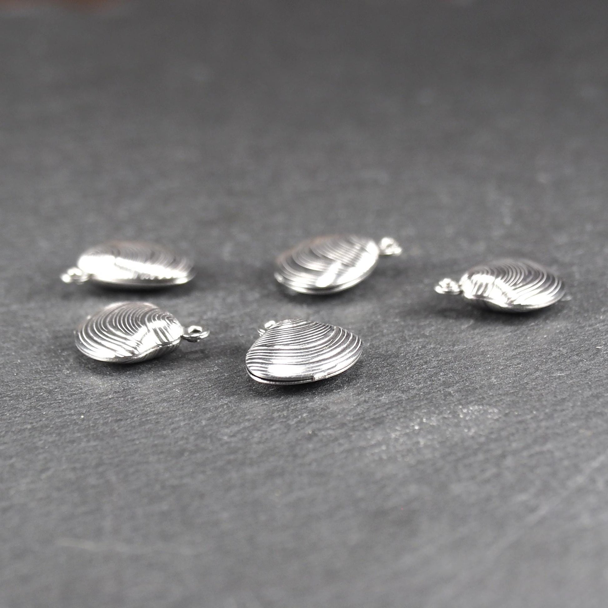 Muschelförmiger silberfarbener Anhänger mit polierter, glänzender Oberfläche, konzentrisch geriffelten Rillen und integrierter runder Öse oben.