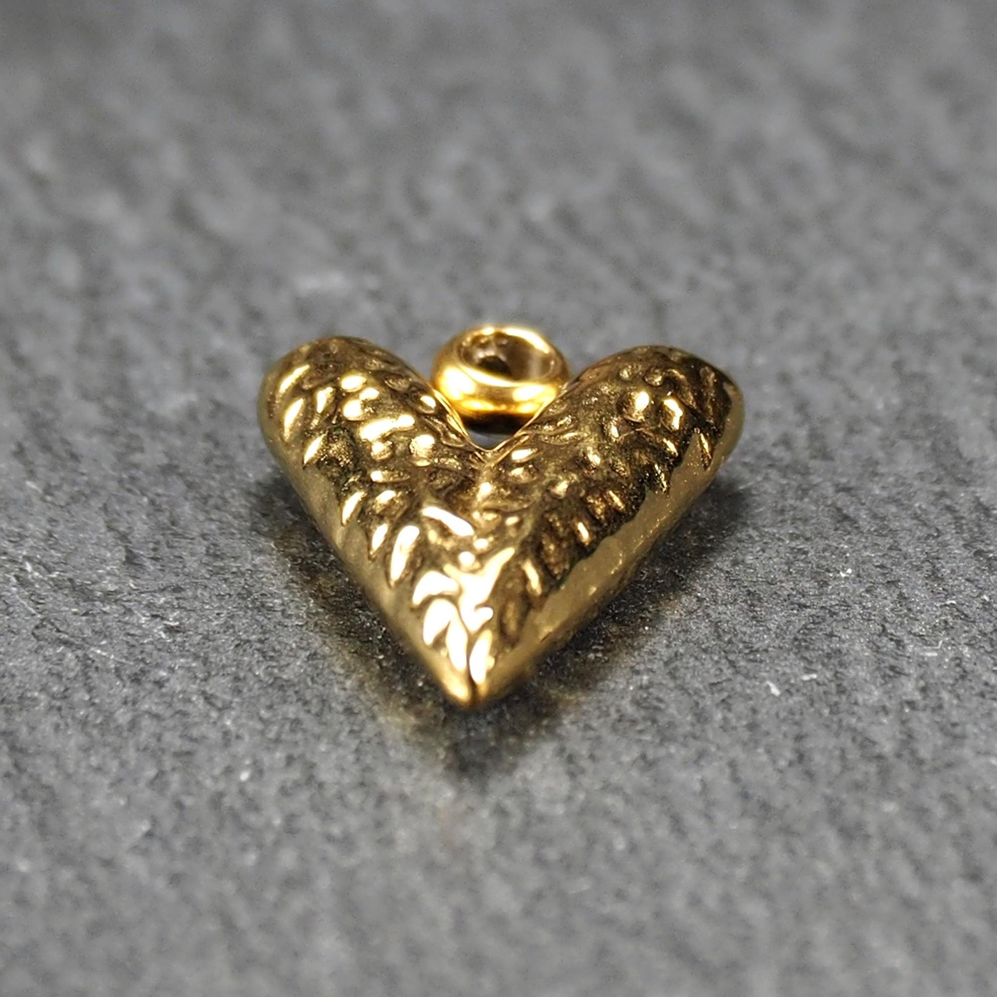 Herzförmiger, vergoldeter Anhänger mit strukturierter Reliefoberfläche, spitz zulaufender Form und integrierter Öse oben.