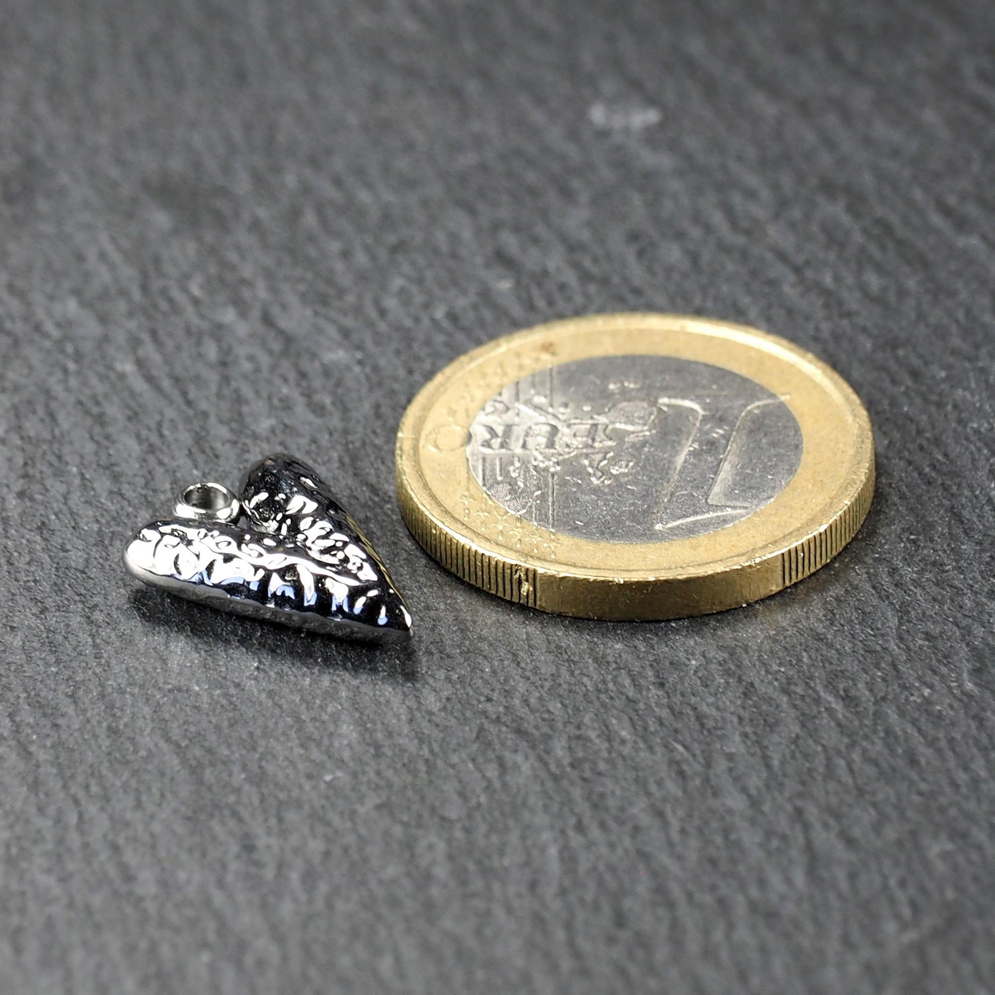 Silberner, hochglanzpolierter herzförmiger Anhänger mit reliefartiger Oberfläche und integrierter Öse, neben einer Ein-Euro-Münze zur Größendarstellung.