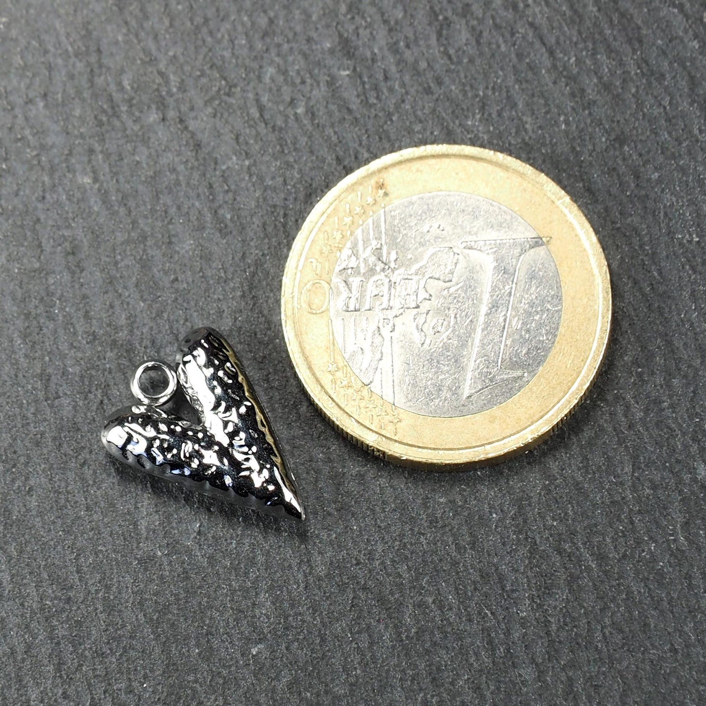 Silberner, hochglanzpolierter herzförmiger Anhänger mit reliefartiger Oberfläche und integrierter Öse, neben einer Ein-Euro-Münze zur Größendarstellung.