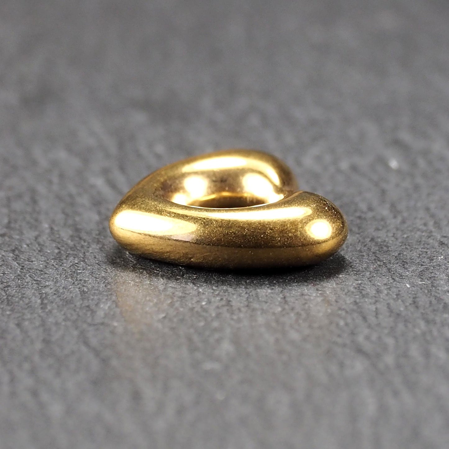 Herzförmiger, goldfarbener Anhänger mit hochglänzender, glatter Oberfläche, rundem durchbrochenem Zentrum und sichtbarer Bohrung.