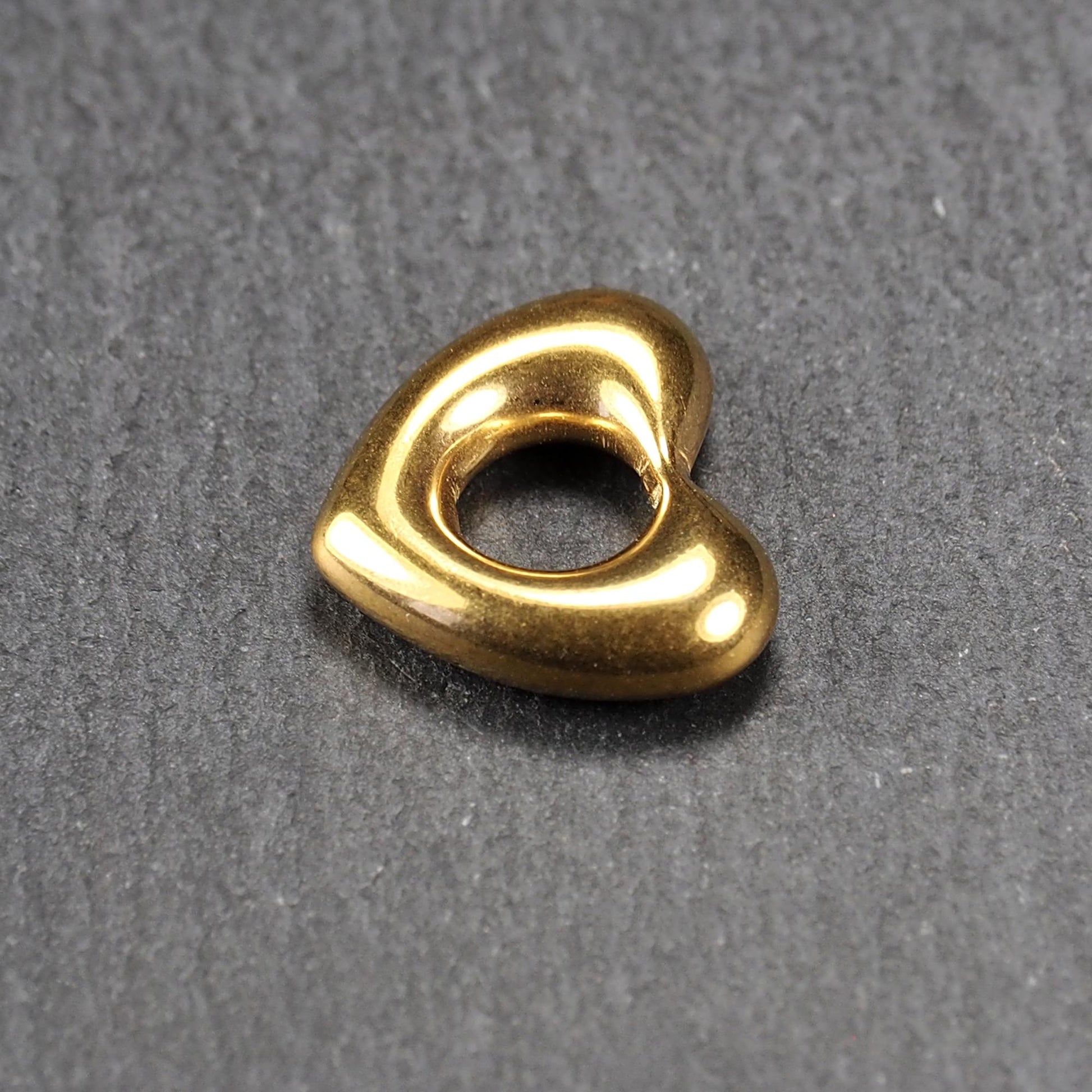 Herzförmiger, goldfarbener Anhänger mit hochglänzender, glatter Oberfläche, rundem durchbrochenem Zentrum und sichtbarer Bohrung.