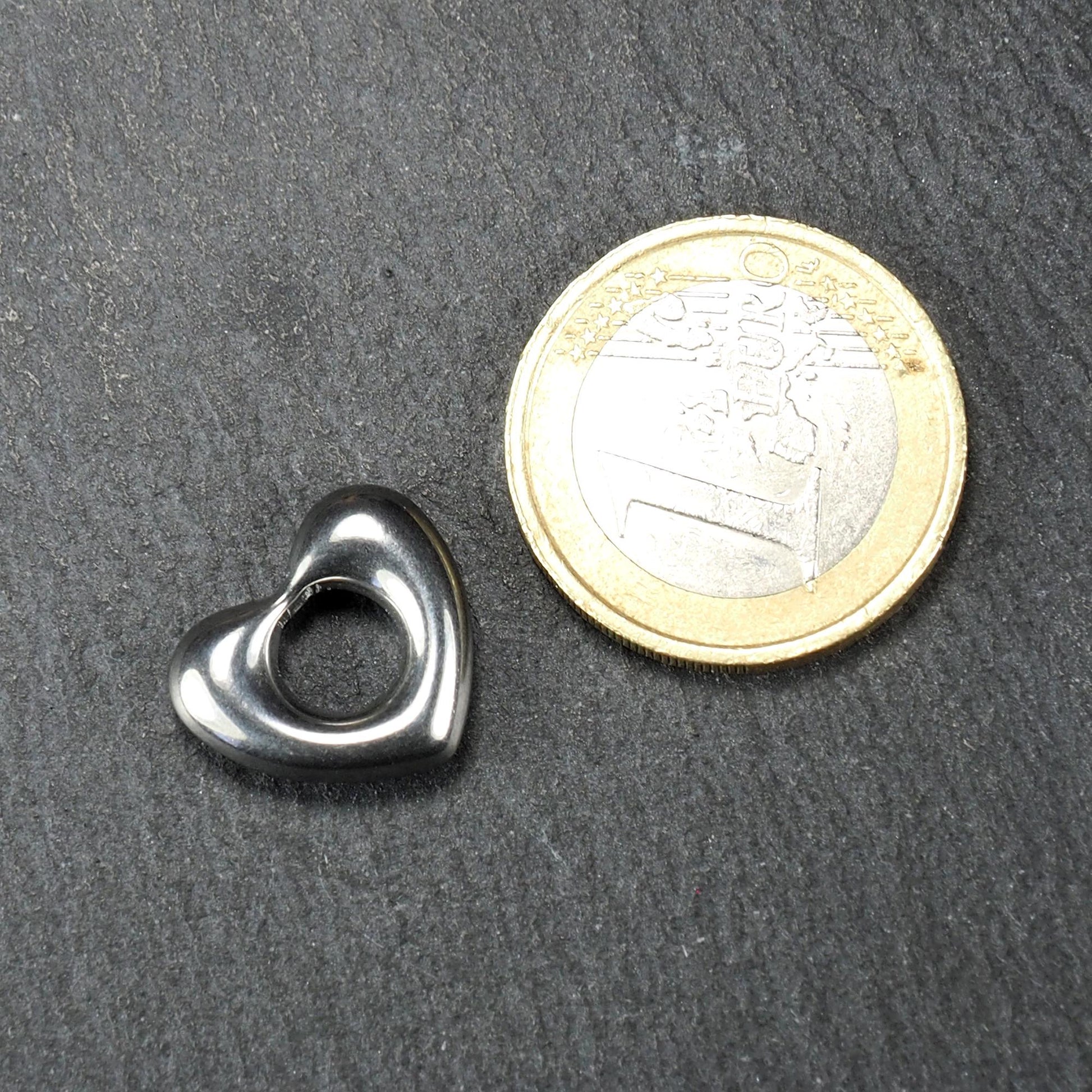 Herzanhänger aus glänzend poliertem Edelstahl mit offenem, durchbrochenem Innenfeld und abgerundeten Kanten, neben einer 1-Euro-Münze.