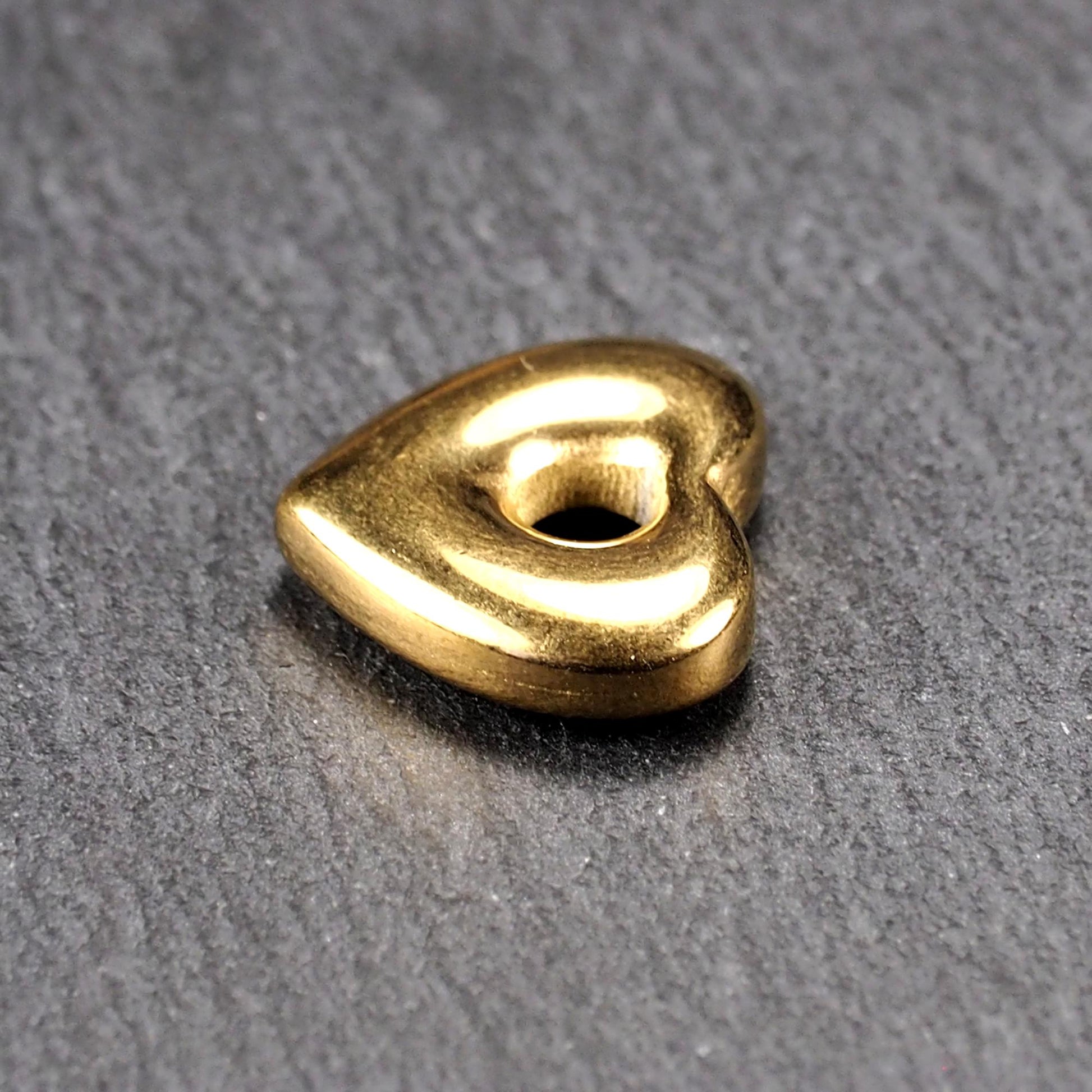 Goldfarbenes, hochglänzendes, abgerundetes Herz mit rundem mittigem Loch, glatte Oberfläche, auf dunkel strukturiertem Untergrund.