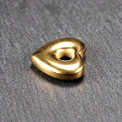 Goldfarbenes, hochglänzendes, abgerundetes Herz mit rundem mittigem Loch, glatte Oberfläche, auf dunkel strukturiertem Untergrund.