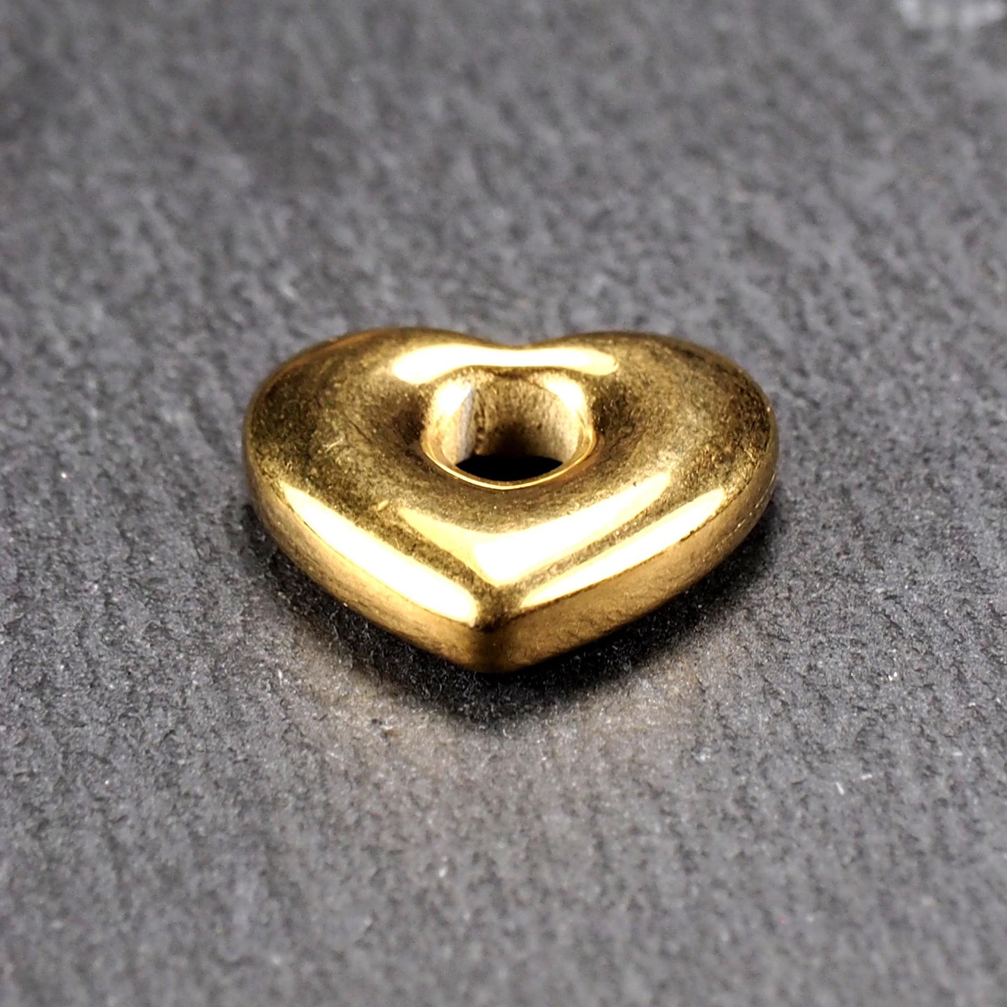 Goldfarbenes, hochglänzendes, abgerundetes Herz mit rundem mittigem Loch, glatte Oberfläche, auf dunkel strukturiertem Untergrund.