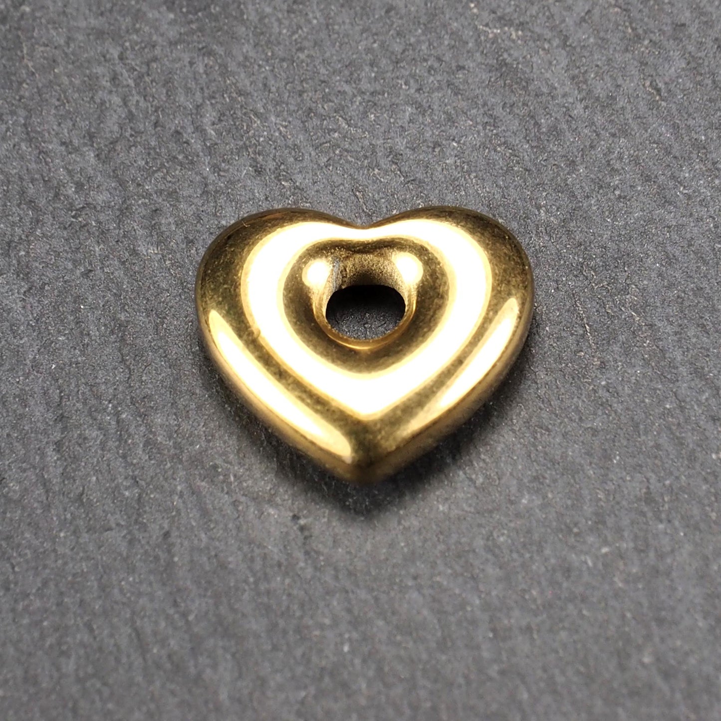 Goldfarbenes, hochglänzendes, abgerundetes Herz mit rundem mittigem Loch, glatte Oberfläche, auf dunkel strukturiertem Untergrund.