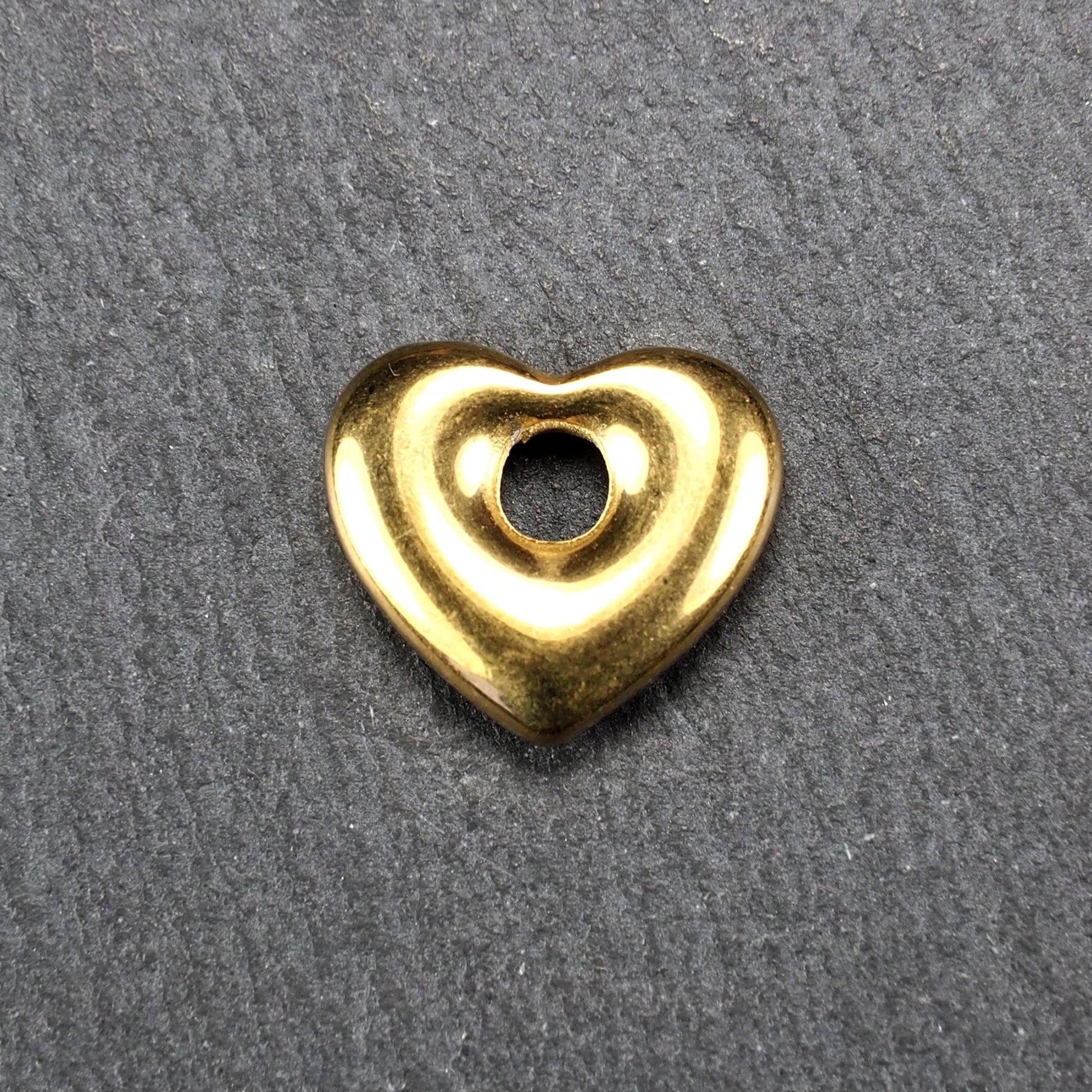 Goldfarbenes, hochglänzendes, abgerundetes Herz mit rundem mittigem Loch, glatte Oberfläche, auf dunkel strukturiertem Untergrund.