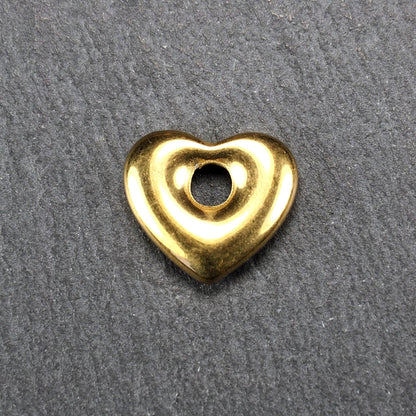 Goldfarbenes, hochglänzendes, abgerundetes Herz mit rundem mittigem Loch, glatte Oberfläche, auf dunkel strukturiertem Untergrund.