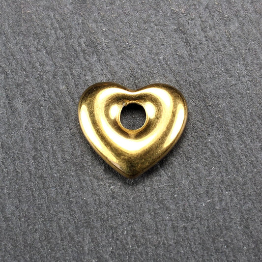 Goldfarbenes, hochglänzendes, abgerundetes Herz mit rundem mittigem Loch, glatte Oberfläche, auf dunkel strukturiertem Untergrund.