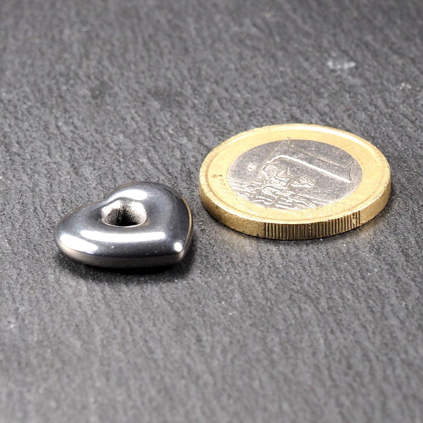 Herzförmiger Anhänger, metallisch dunkelgrau, glänzend poliert, mit rundem mittigem Fädelloch; daneben eine Ein-Euro-Münze zum Größenvergleich.