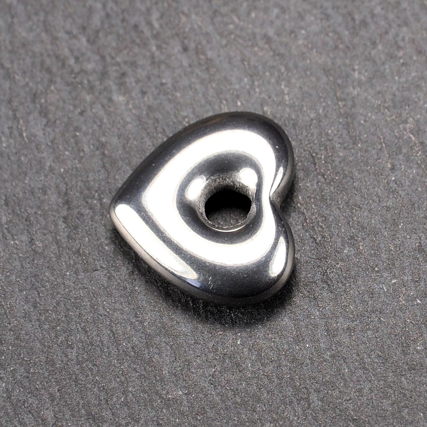 Herzförmiger, silberfarben glänzender Metallanhänger mit glatter Oberfläche und mittigem rundem Fädelloch.