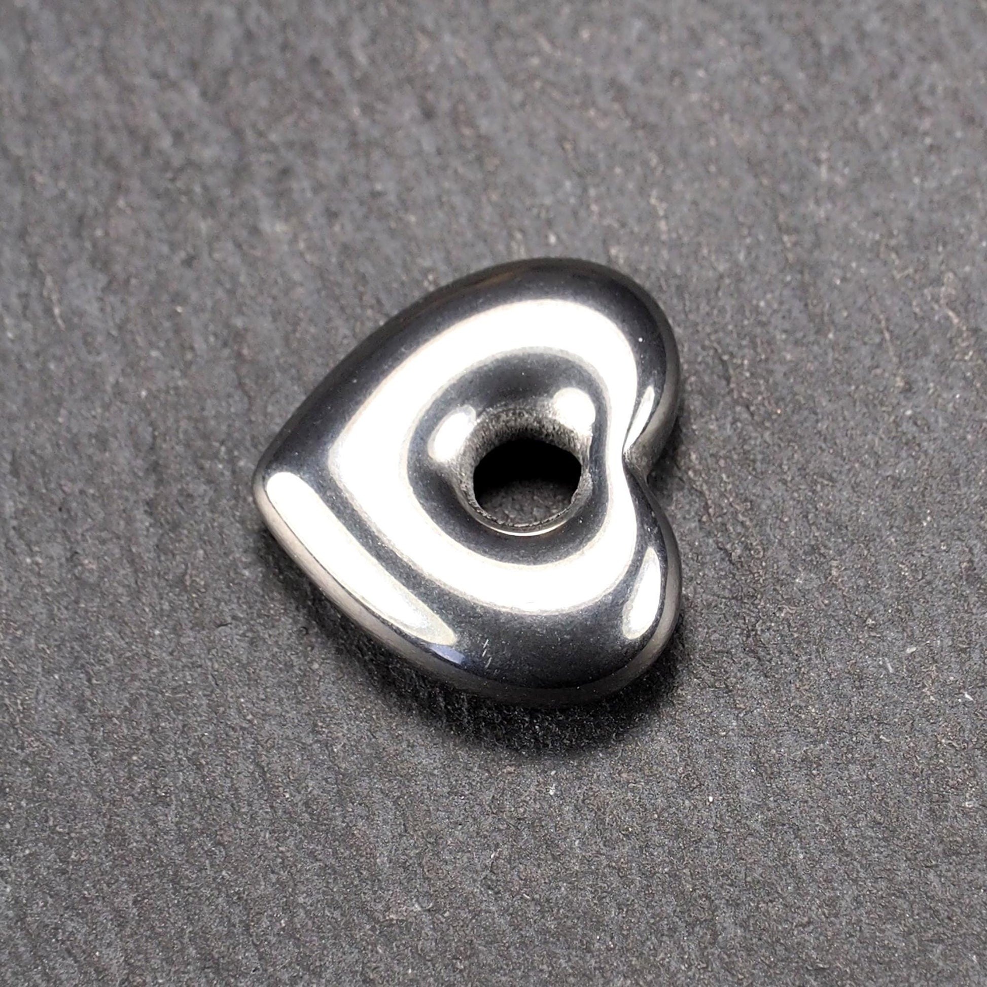 Herzförmiger, silberfarben glänzender Metallanhänger mit glatter Oberfläche und mittigem rundem Fädelloch.