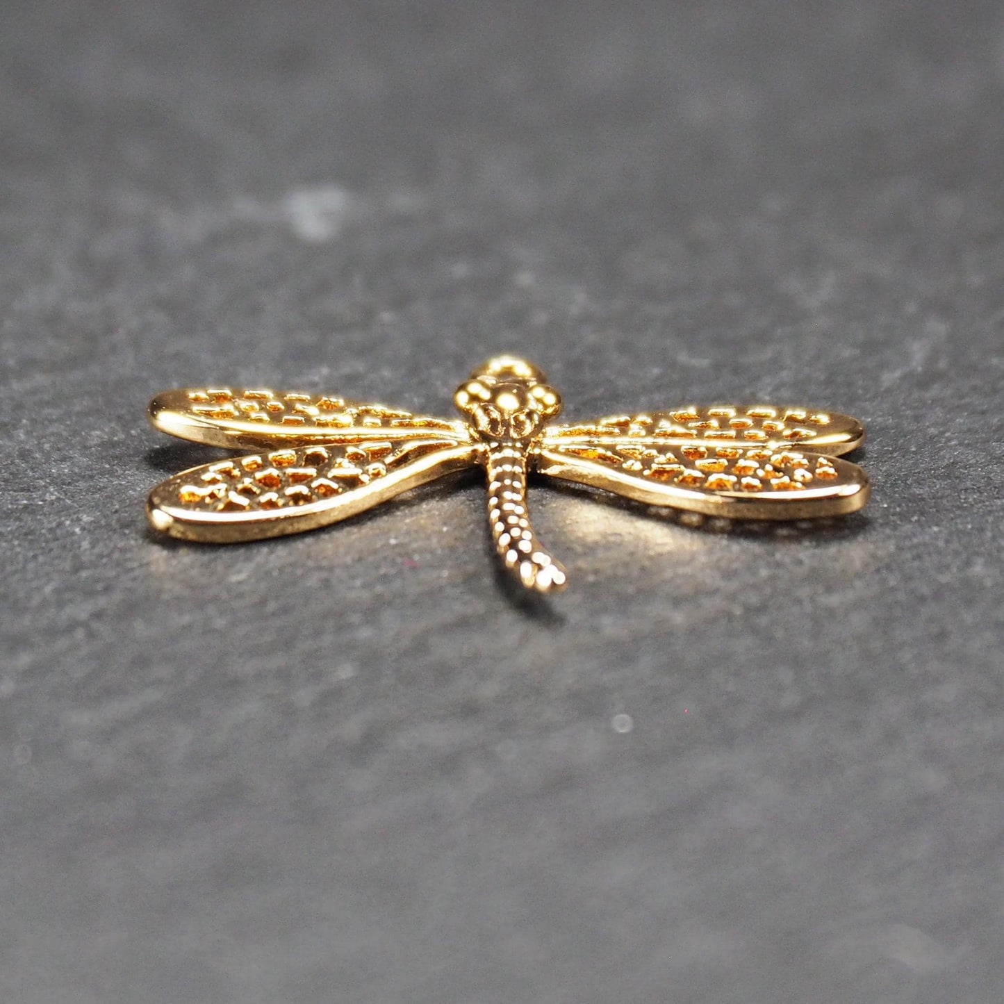 Libellen-Anhänger aus goldfarbenem, glänzendem Metall mit durchbrochenen, netzartigen Flügeln, schlankem segmentiertem Körper und kleiner runder Aufhängöse.