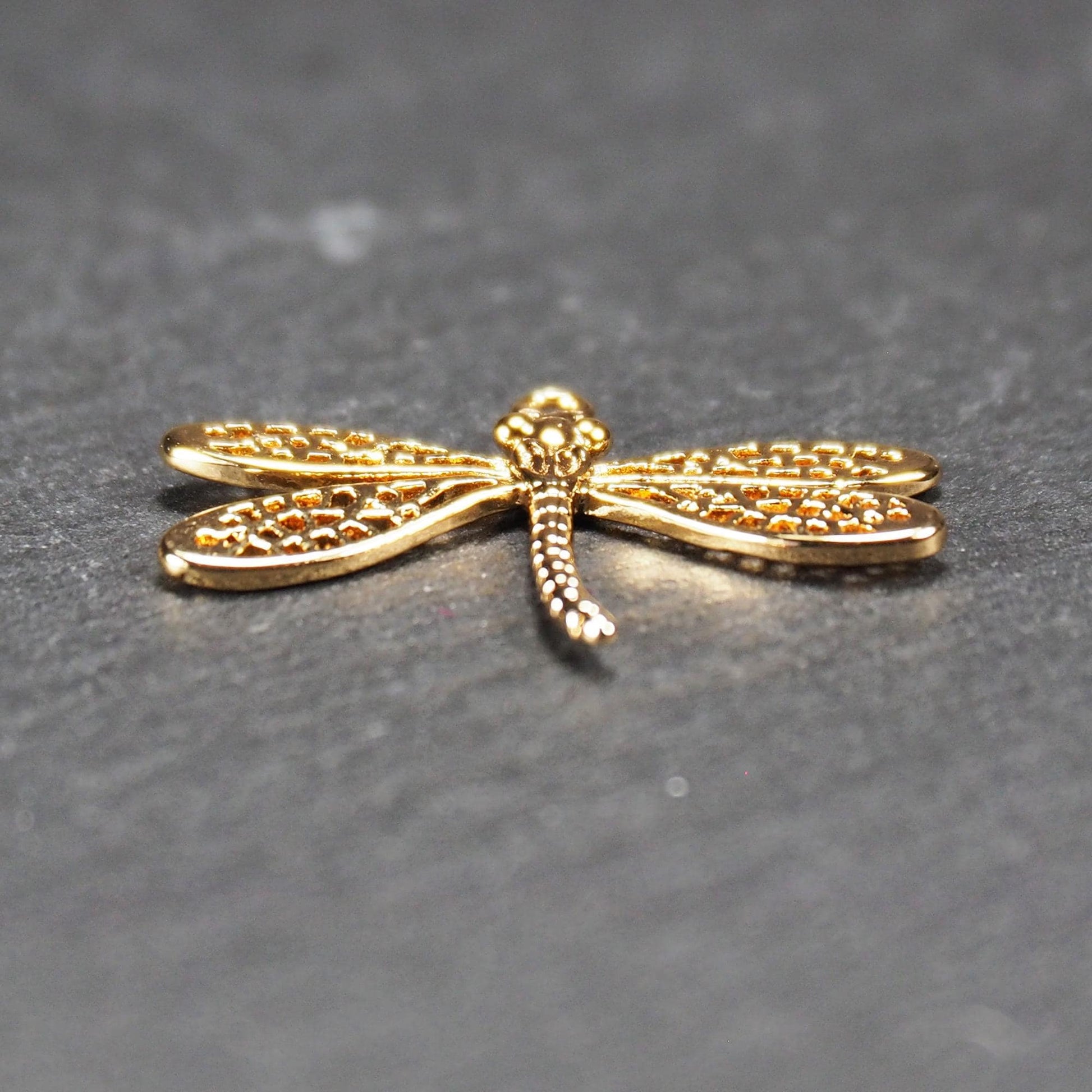 Libellen-Anhänger aus goldfarbenem, glänzendem Metall mit durchbrochenen, netzartigen Flügeln, schlankem segmentiertem Körper und kleiner runder Aufhängöse.
