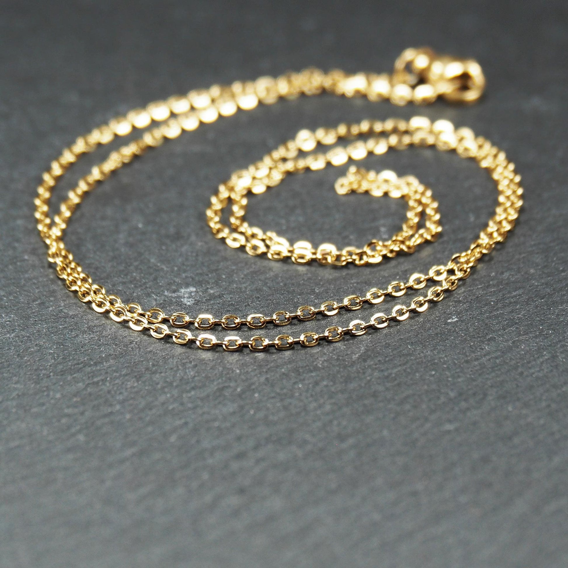 Feine goldfarbene Ankerkette mit glänzender Oberfläche, filigranen ovalen Gliedern in Spiralform und sichtbarem Karabinerverschluss.