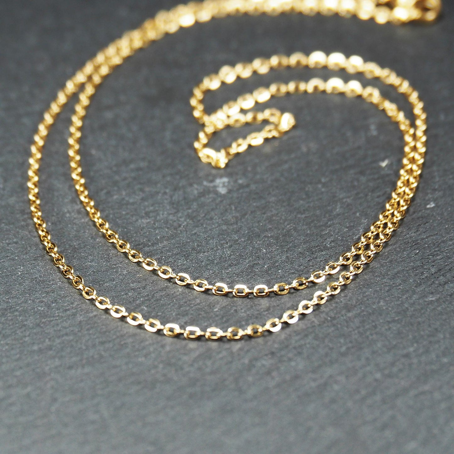 Feine goldfarbene Ankerkette mit glänzender Oberfläche, filigranen ovalen Gliedern in Spiralform und sichtbarem Karabinerverschluss.