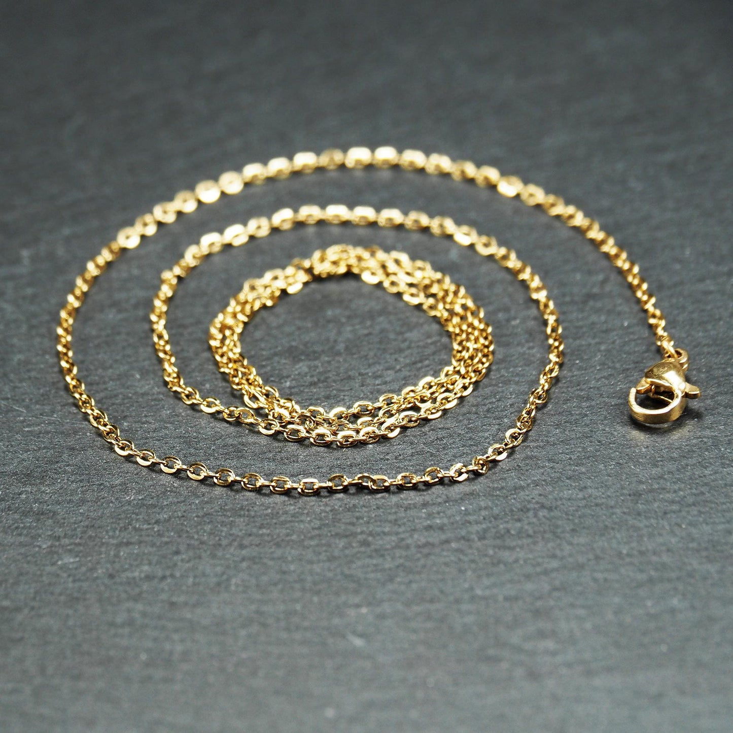 Feine goldfarbene Ankerkette mit glänzender Oberfläche, filigranen ovalen Gliedern in Spiralform und sichtbarem Karabinerverschluss.