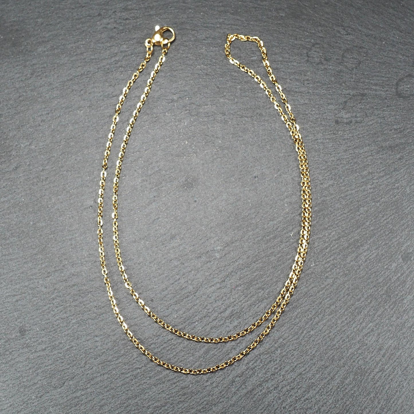 Feine goldfarbene Ankerkette mit glänzender Oberfläche, filigranen ovalen Gliedern in Spiralform und sichtbarem Karabinerverschluss.