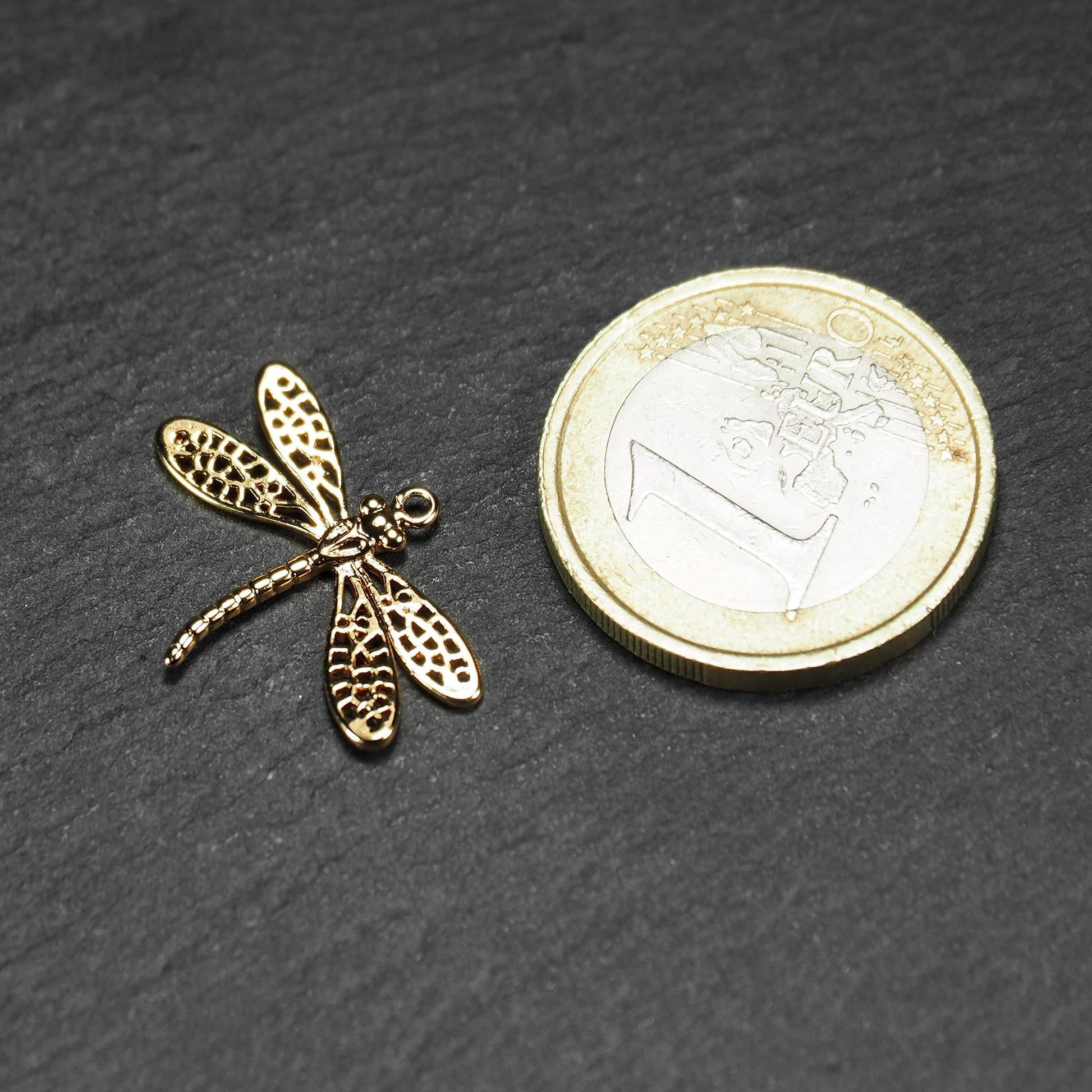 Libellen-Anhänger aus goldfarbenem, glänzendem Metall mit durchbrochenen, netzartigen Flügeln, schlankem segmentiertem Körper und kleiner runder Aufhängöse. Neben einer 1 Euro Münze zum Größenvergleich.