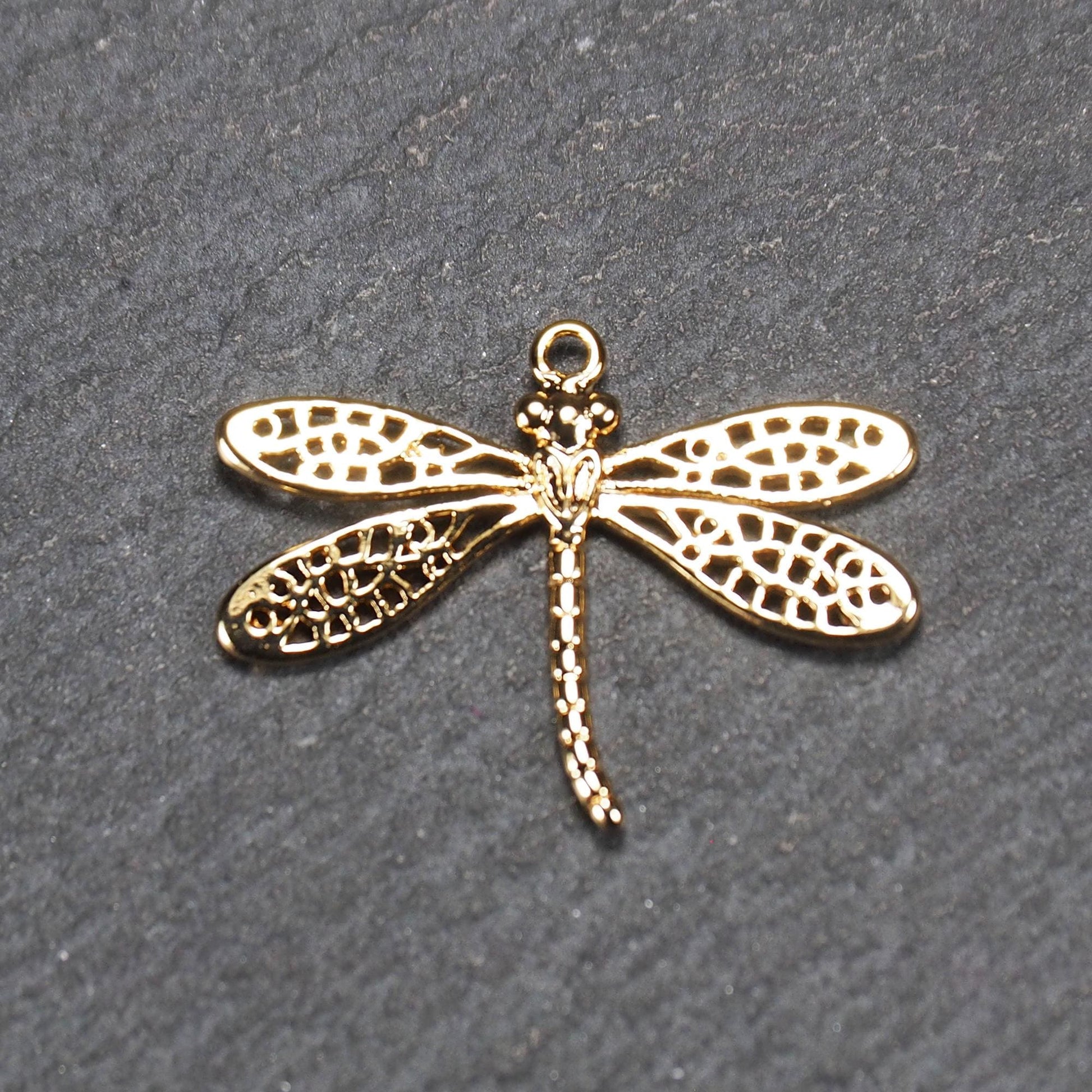 Libellen-Anhänger aus goldfarbenem, glänzendem Metall mit durchbrochenen, netzartigen Flügeln, schlankem segmentiertem Körper und kleiner runder Aufhängöse.