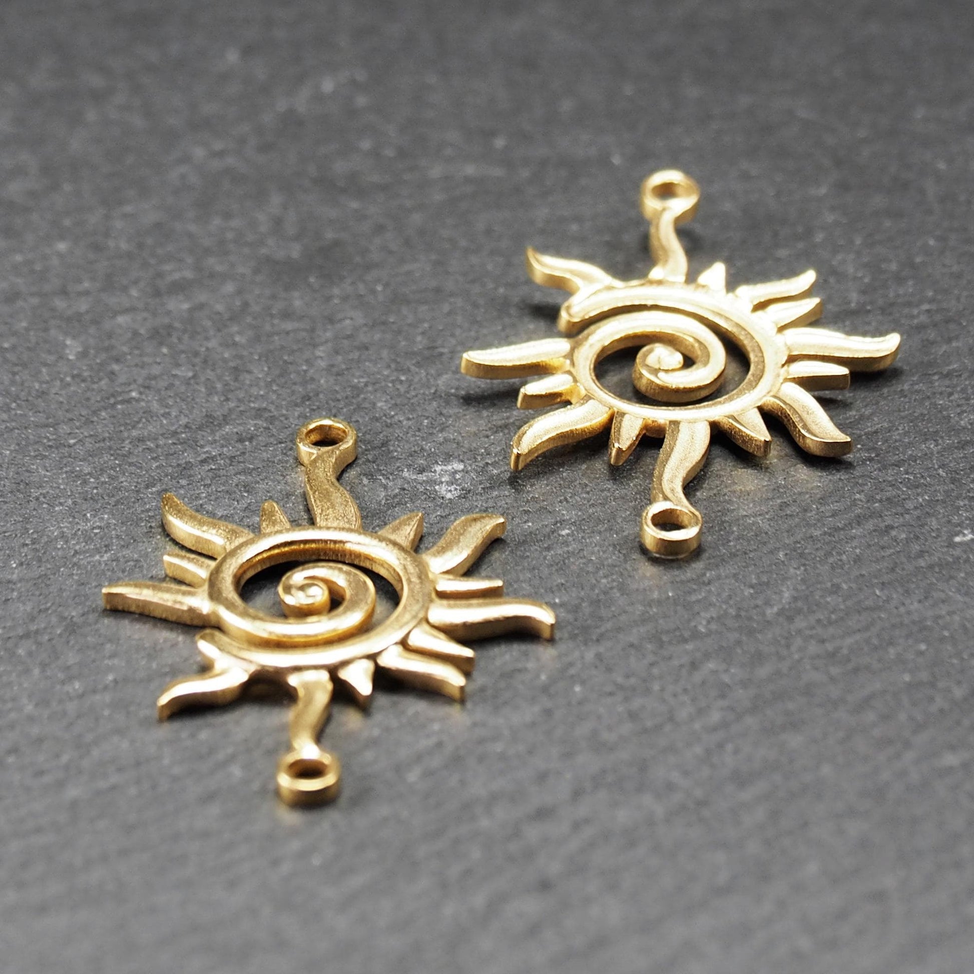 Goldfarbener, durchbrochener Sonnen-Verbinder mit spiralförmigem Zentrum, unregelmäßigen wellenförmigen Strahlen und je einer Öse oben und unten.  Vorder- und Rückseite.