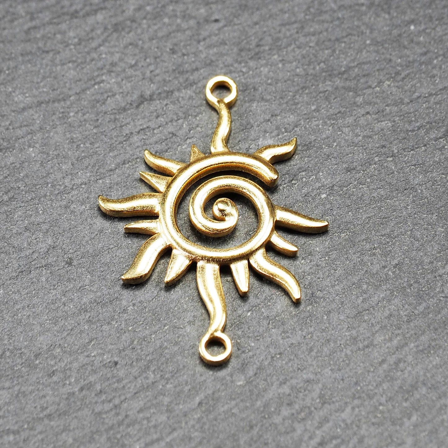 Goldfarbener, durchbrochener Sonnen-Verbinder mit spiralförmigem Zentrum, unregelmäßigen wellenförmigen Strahlen und je einer Öse oben und unten.