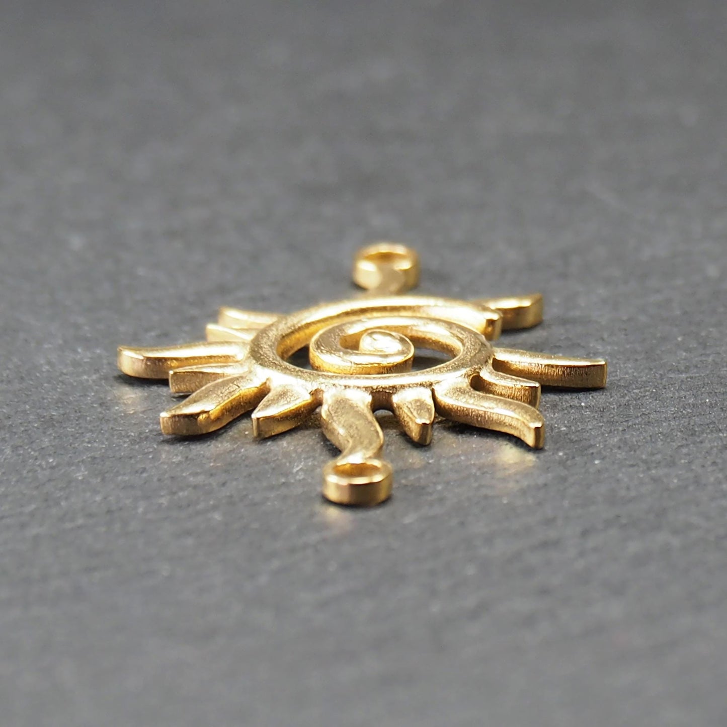 Goldfarbener, durchbrochener Sonnen-Verbinder mit spiralförmigem Zentrum, unregelmäßigen wellenförmigen Strahlen und je einer Öse oben und unten.