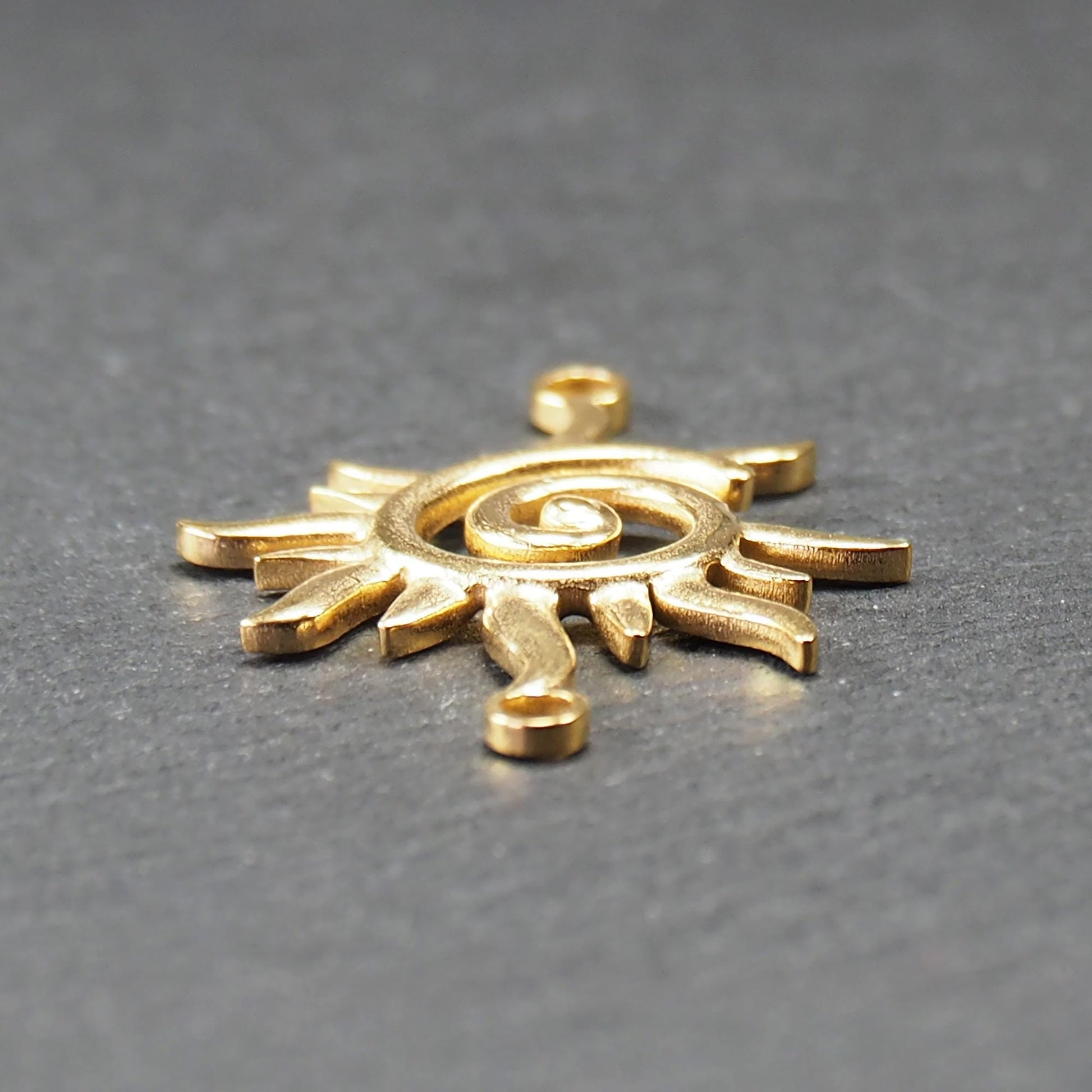 Goldfarbener, durchbrochener Sonnen-Verbinder mit spiralförmigem Zentrum, unregelmäßigen wellenförmigen Strahlen und je einer Öse oben und unten.