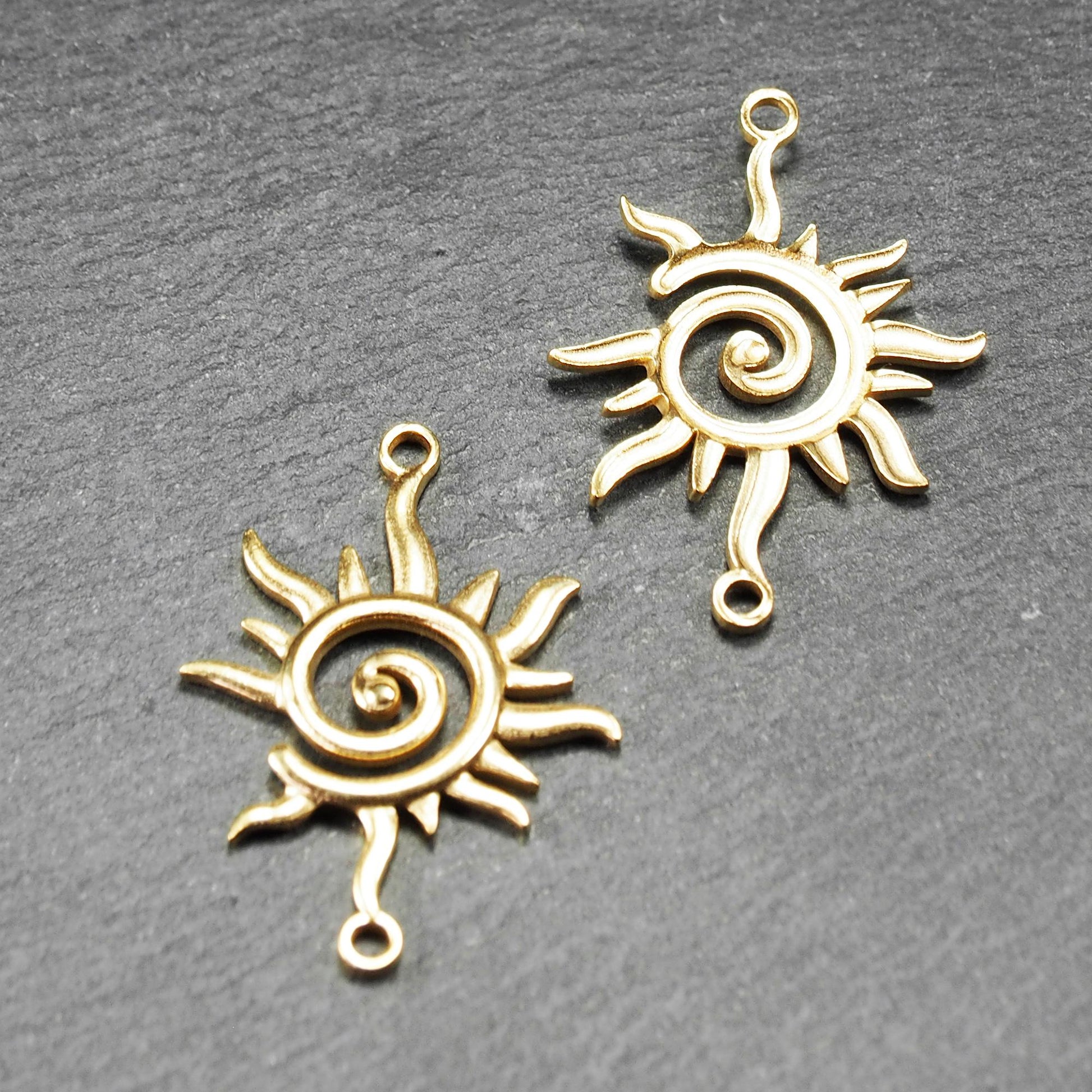 Goldfarbener, durchbrochener Sonnen-Verbinder mit spiralförmigem Zentrum, unregelmäßigen wellenförmigen Strahlen und je einer Öse oben und unten.  Vorder- und Rückseite.