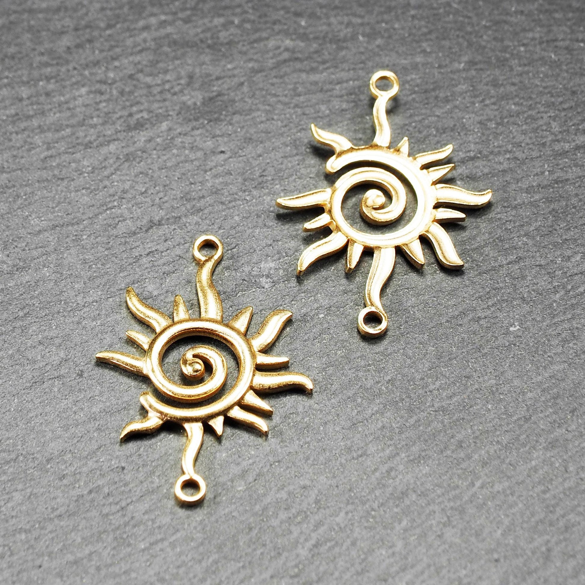 Goldfarbener, durchbrochener Sonnen-Verbinder mit spiralförmigem Zentrum, unregelmäßigen wellenförmigen Strahlen und je einer Öse oben und unten.  Vorder- und Rückseite.
