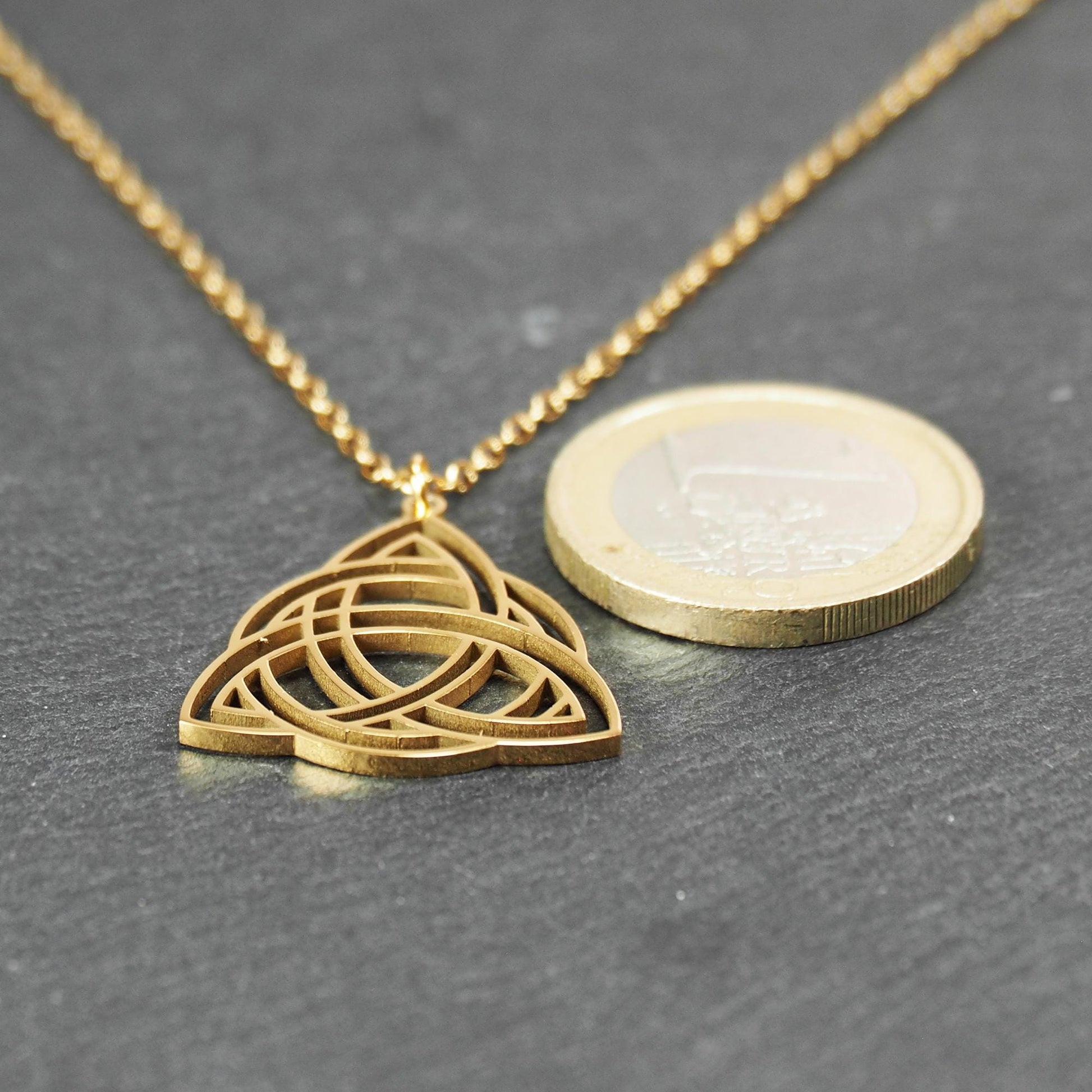 Vergoldeter Anhänger in offener Triquetra-Knotenform mit filigranen Durchbrüchen, an dünner goldfarbener Kette und sichtbarer Öse. 1-Euro-Münze als Größenvergleich.