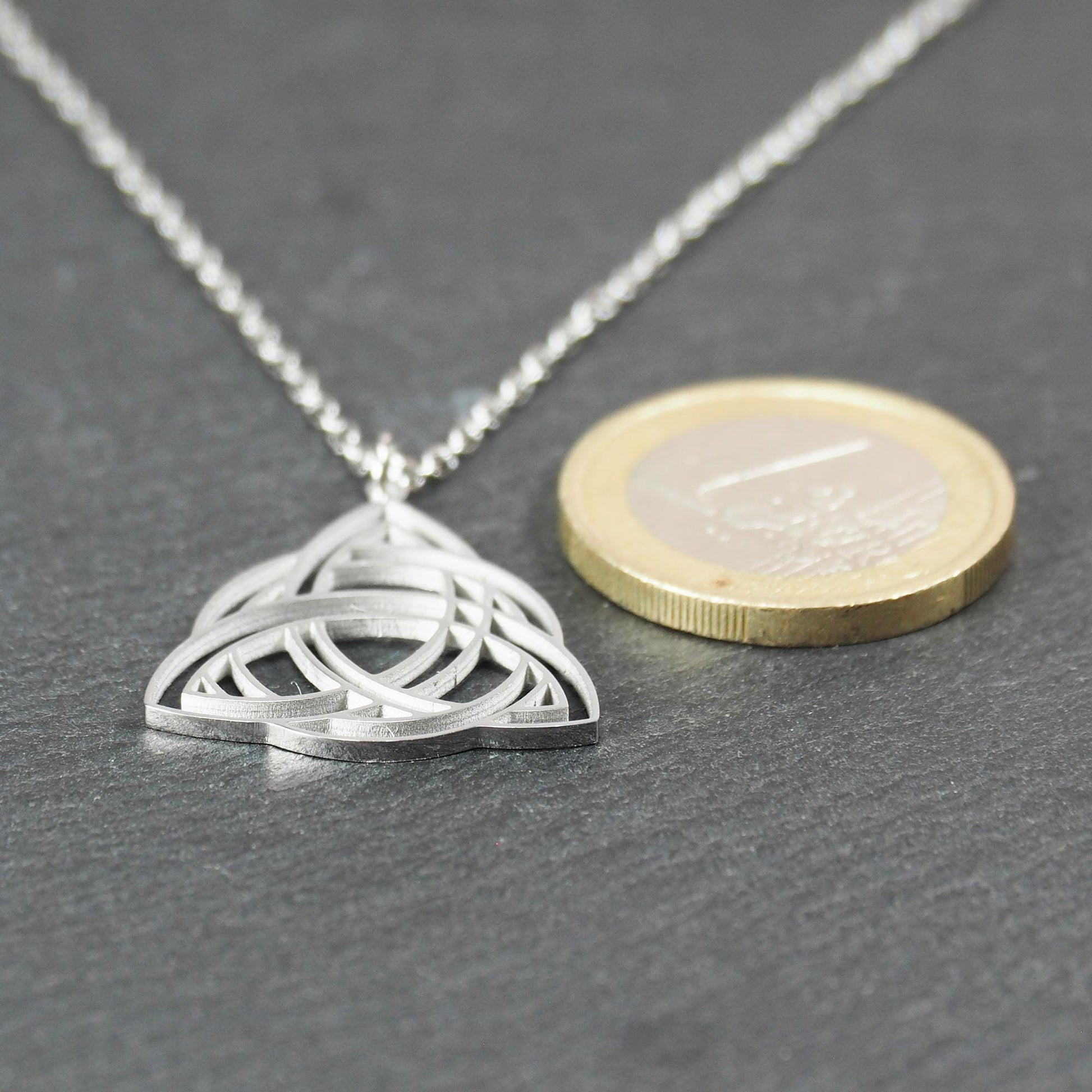 Anhänger, silberfarben glänzend; dreieckige, offene Triquetra-Knotenform mit filigraner Durchbruchstruktur und sichtbarer Öse an feiner Gliederkette. 1-Euro-Münze als Größenvergleich.