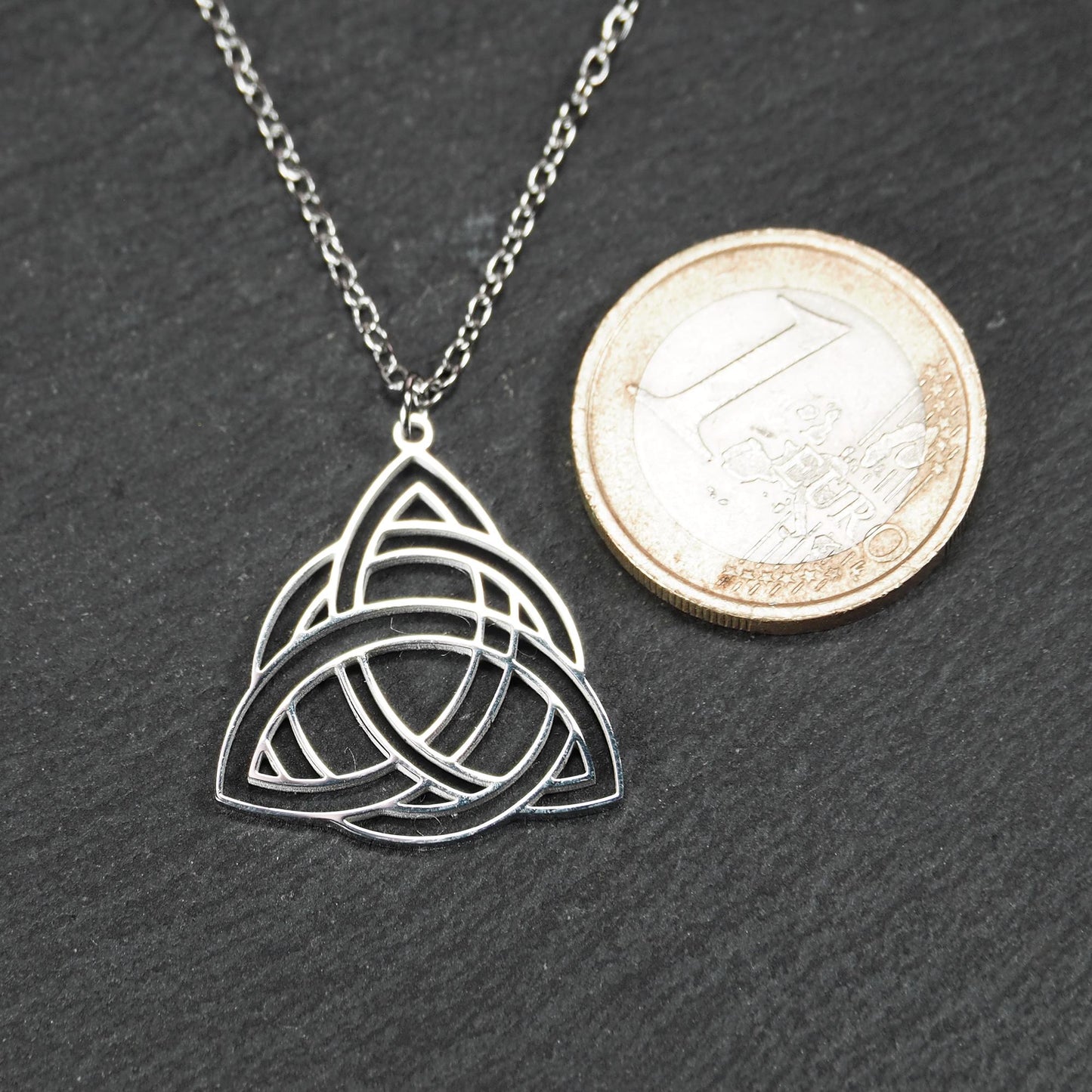 Anhänger, silberfarben glänzend; dreieckige, offene Triquetra-Knotenform mit filigraner Durchbruchstruktur und sichtbarer Öse an feiner Gliederkette. 1-Euro-Münze als Größenvergleich.