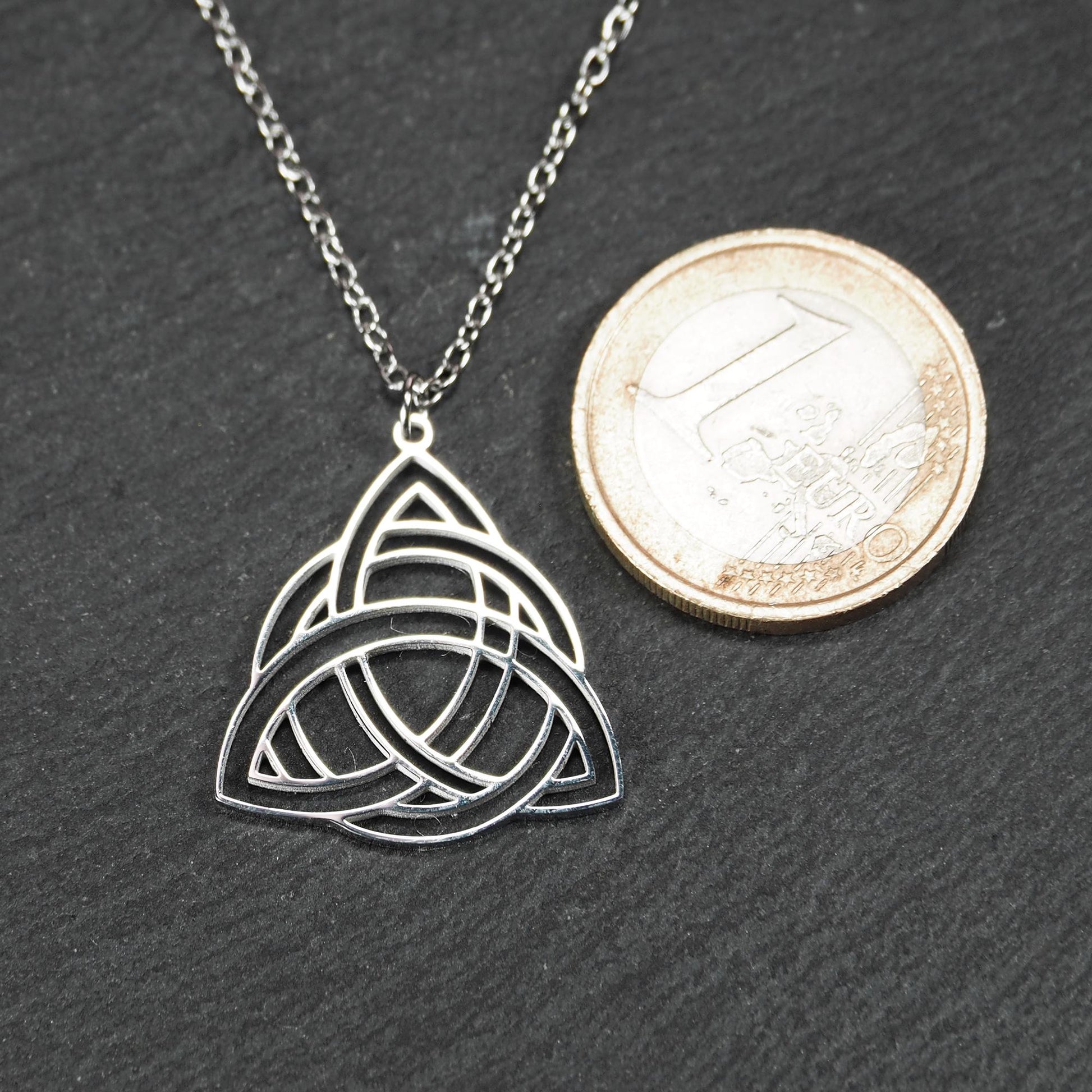 Anhänger, silberfarben glänzend; dreieckige, offene Triquetra-Knotenform mit filigraner Durchbruchstruktur und sichtbarer Öse an feiner Gliederkette. 1-Euro-Münze als Größenvergleich.