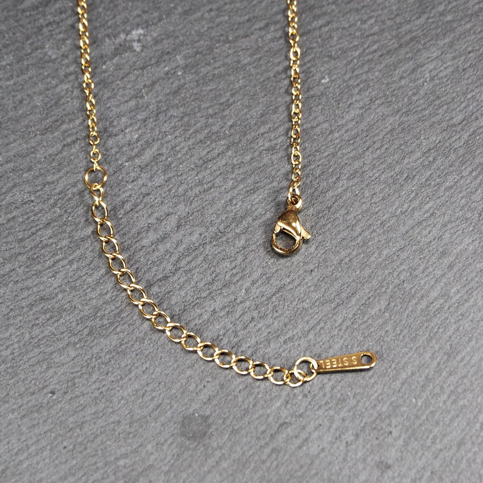 Feine goldfarbene Gliederkette mit Karabinerverschluss, Verlängerungskettchen und kleinem rechteckigen Markenplättchen.