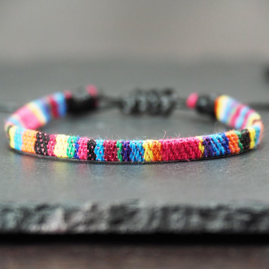 Gewebtes, flaches Armband mit regenbogenfarbenen Streifen, schmaler Form und feiner Webstruktur, schwarzer Schiebeknotenverschluss.